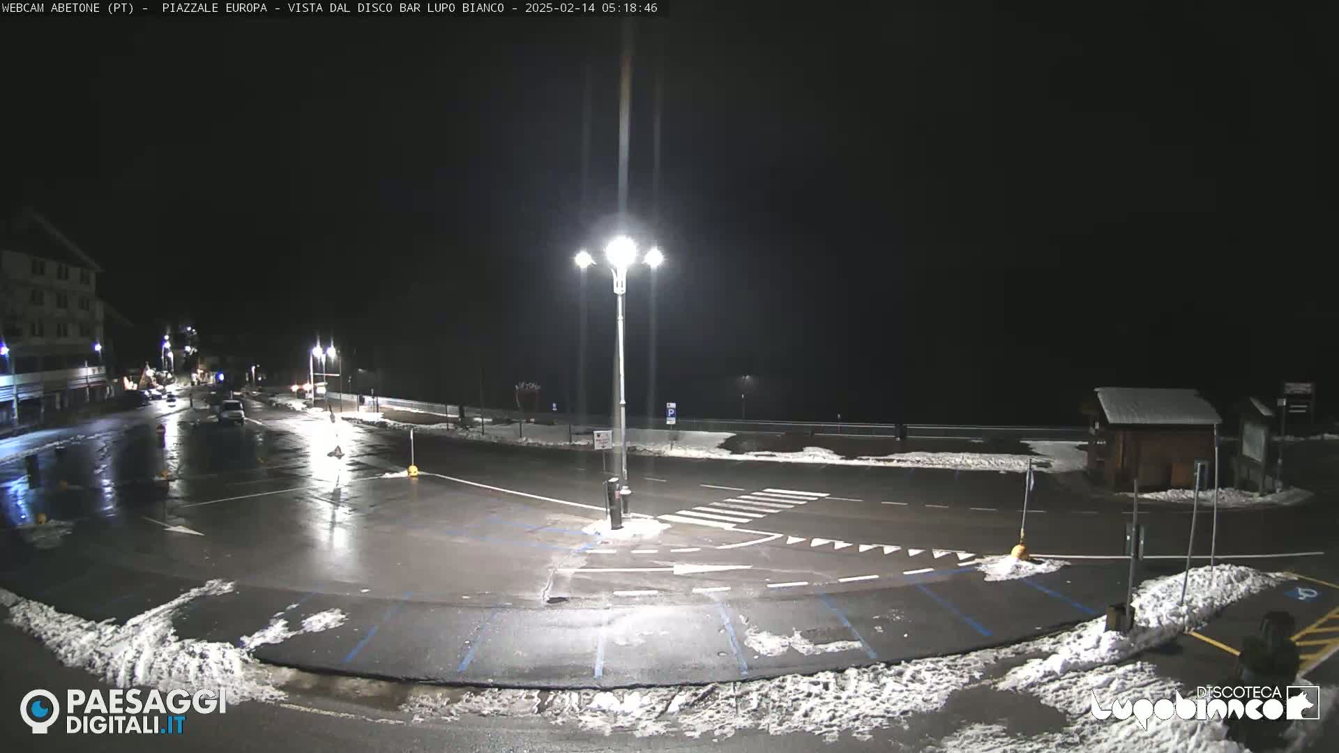A mostly empty, wet parking lot at night with some snow on the ground and illuminated by streetlights.
