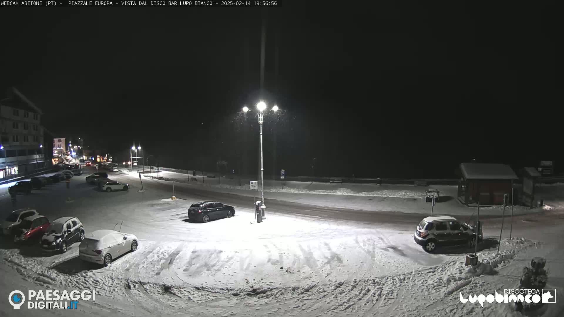 A snow-covered parking lot and road at night, with several cars parked, and light snowfall.