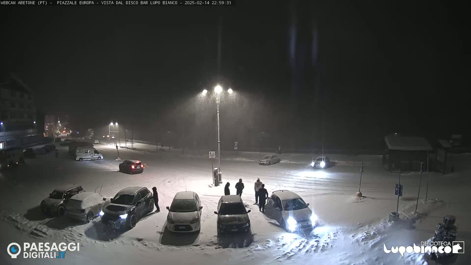 A snow-covered parking lot at night, illuminated by streetlights, with several cars parked and a few people standing around.