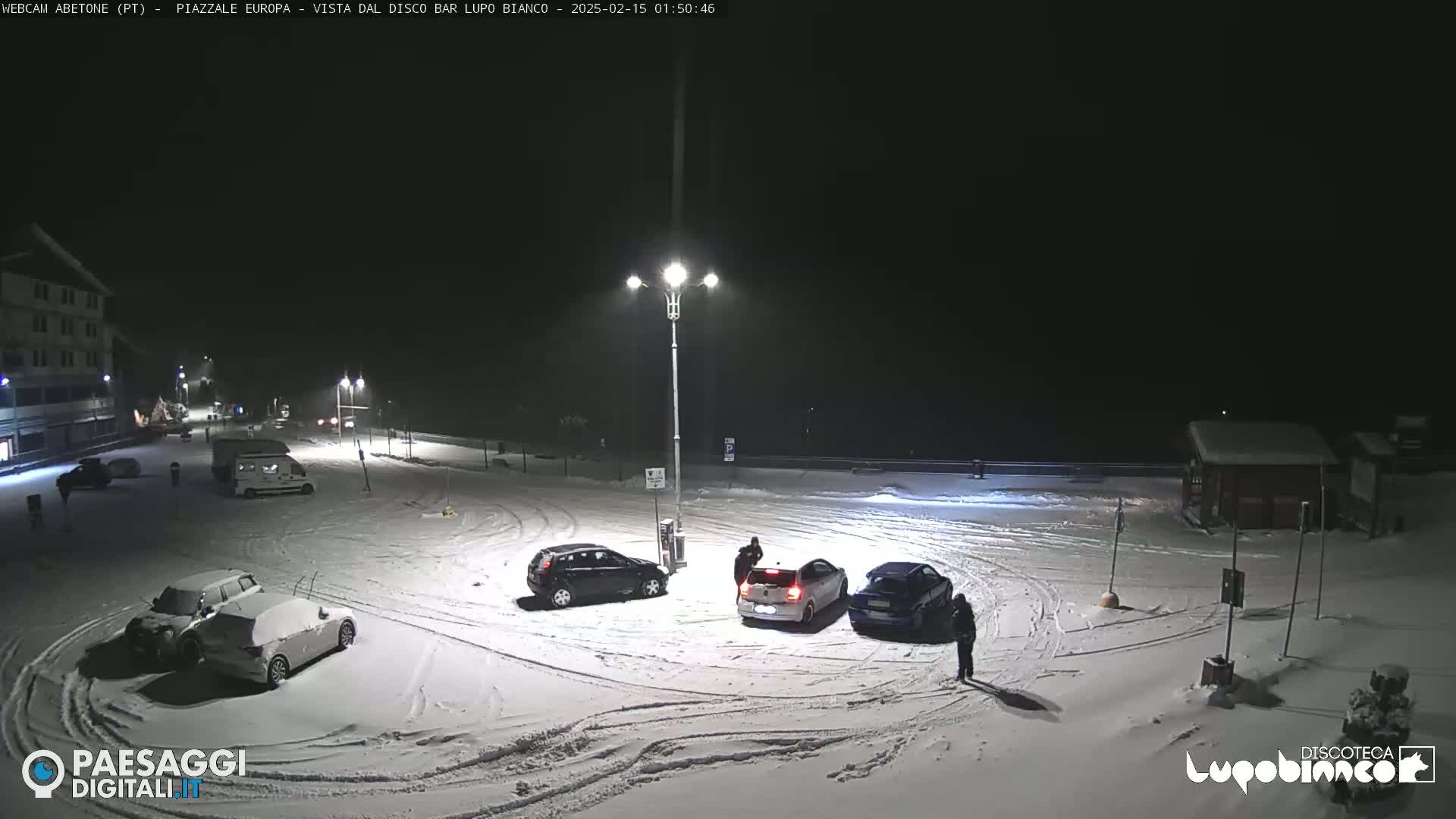 A snow-covered parking lot at night, illuminated by streetlights, with several cars parked and a few people standing near them.