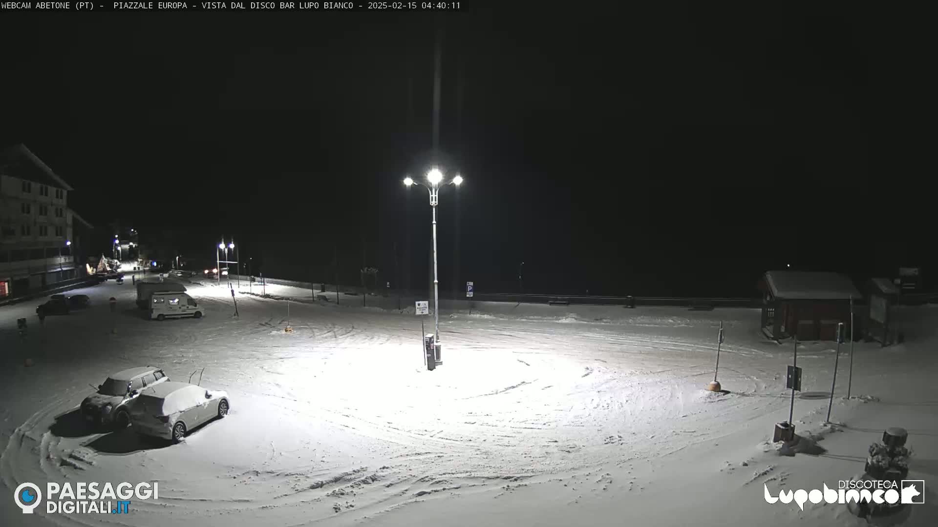 A snow-covered parking lot is illuminated at night by a tall lamppost, with several vehicles parked and a building visible in the distance.