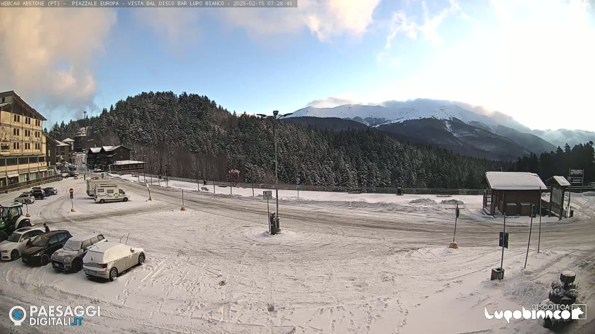 A snow-covered parking lot and road are situated in front of a snow-dusted evergreen forest and a partially snow-capped mountain range under a partly cloudy sky.