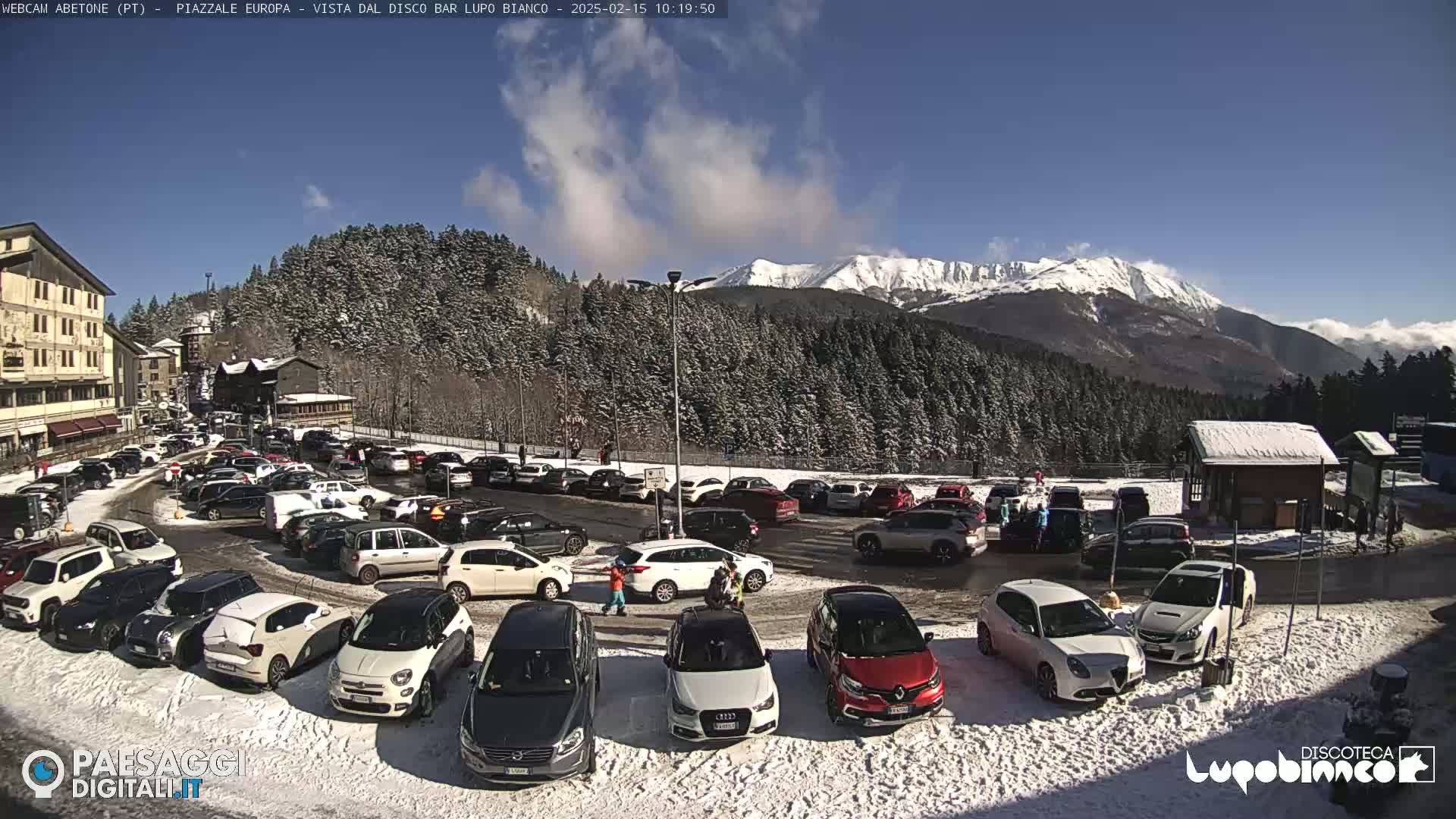 A snow-covered parking lot filled with cars is situated in front of a building and a snow-dusted forest, with a mountain range in the background under a sunny sky.