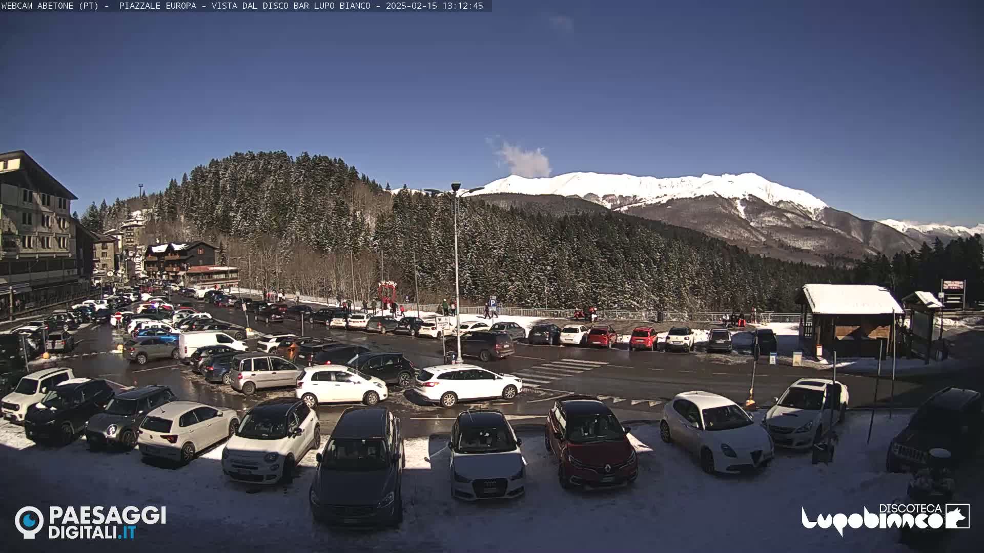 A sunny day shows a snow-dusted mountain range in the background, overlooking a parking lot full of cars near a building, with some people walking around.