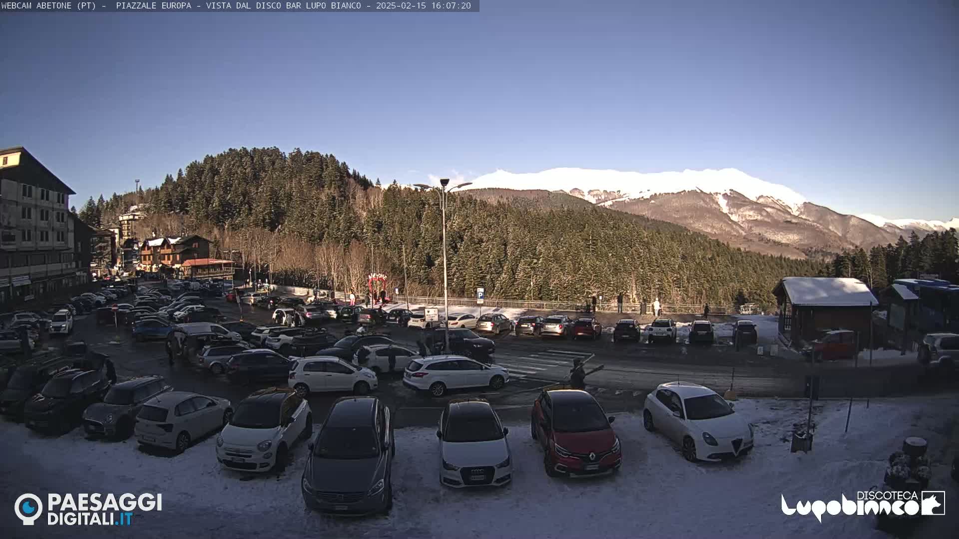 A sunny day reveals a snow-dusted parking lot full of cars, situated in front of a mountain range covered in snow and a forest of dark green trees.
