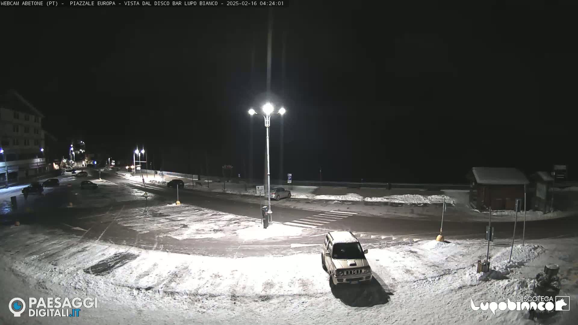 A snow-covered nighttime parking lot and road are illuminated by streetlights, with a few cars visible.