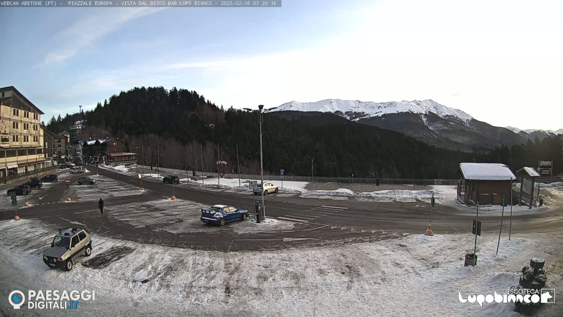 A mostly empty snow-dusted parking lot is situated in front of a building and overlooks a snow-capped mountain range under a mostly clear sky.