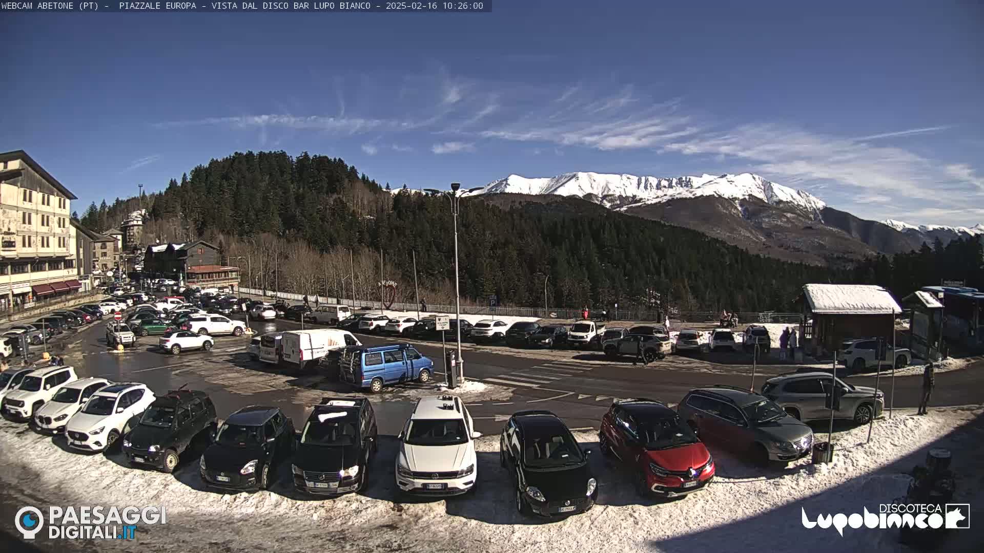 A sunny day reveals a snow-covered mountain range in the background, overlooking a parking lot filled with cars and a few pedestrians near a small building.