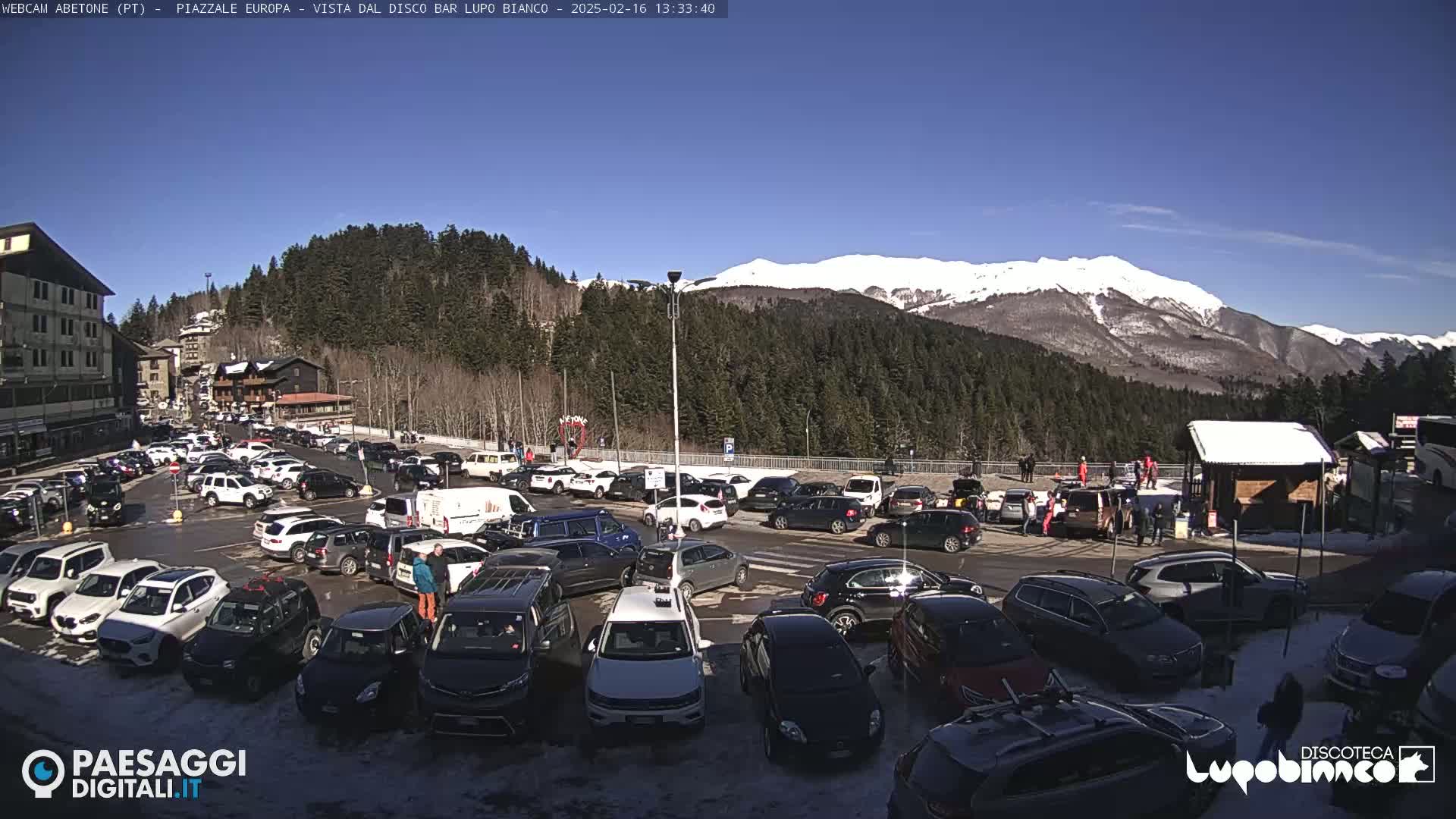 A sunny day in a mountain town parking lot, filled with cars, overlooks snow-capped mountains in the distance.