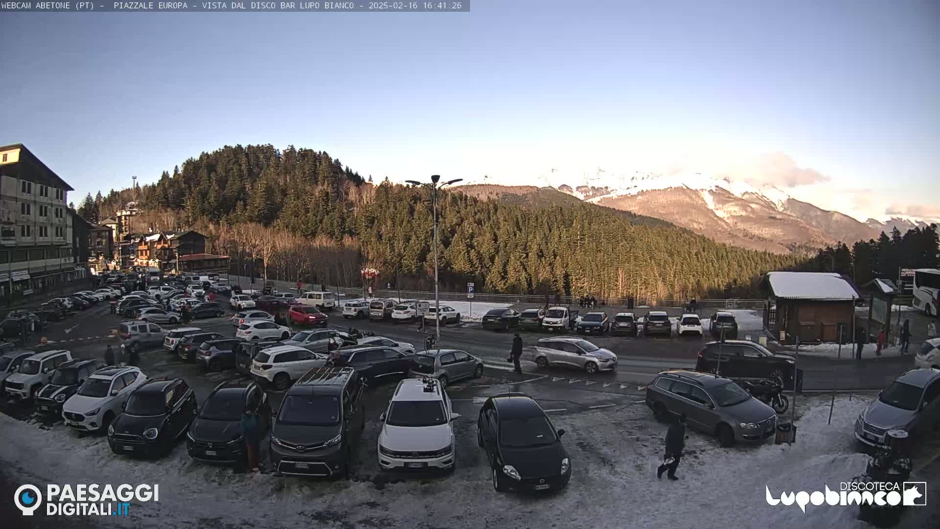A large parking lot filled with cars is situated in front of a building and a snow-covered mountain range under a clear sky.