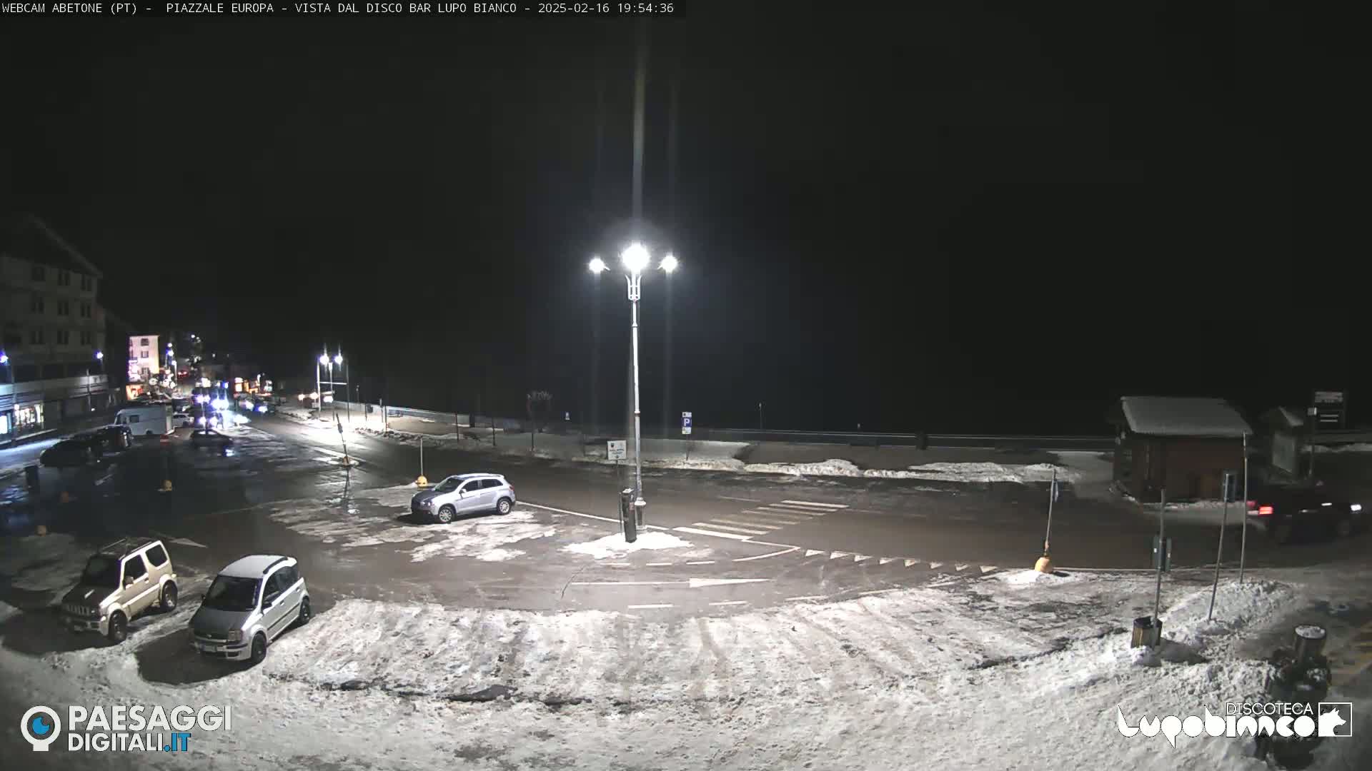 A nighttime view of a snow-dusted parking lot and roadway, with several cars parked and a few driving, under a dark, clear sky.