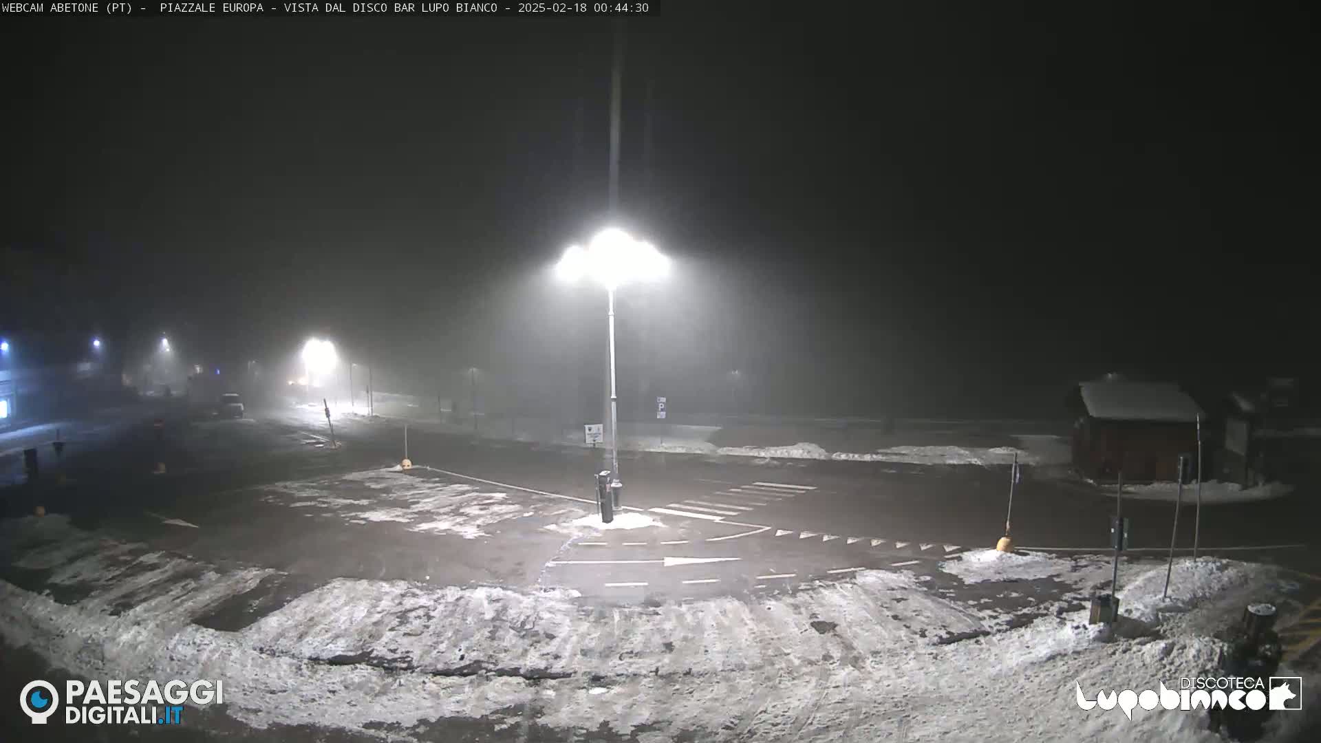 A mostly empty, snow-dusted parking lot is dimly lit at night under foggy conditions.