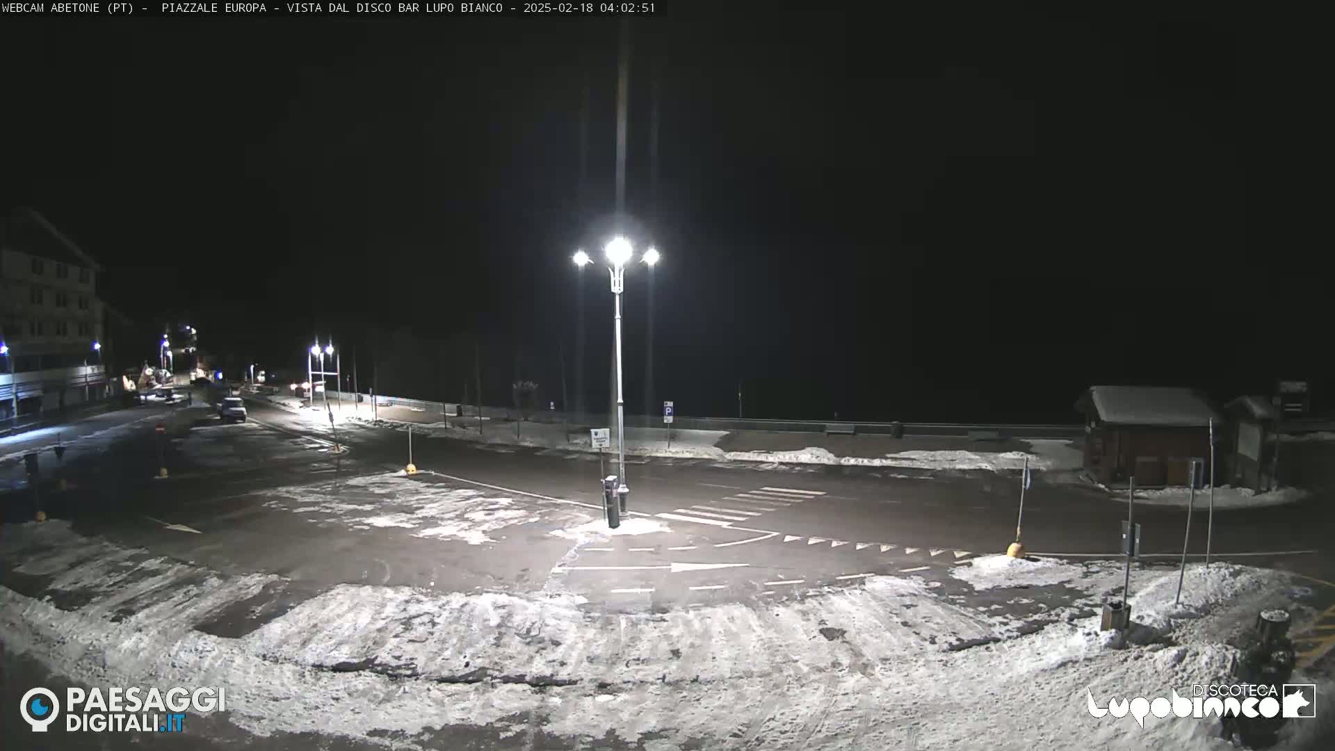 A nighttime view of a snow-covered empty parking lot and street intersection, illuminated by streetlights, with a building visible in the background.