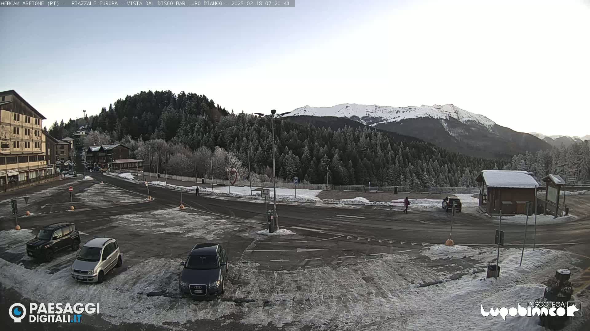 A snow-dusted parking lot with several cars is situated before a line of evergreen trees and a snow-capped mountain range under a clear, light sky.