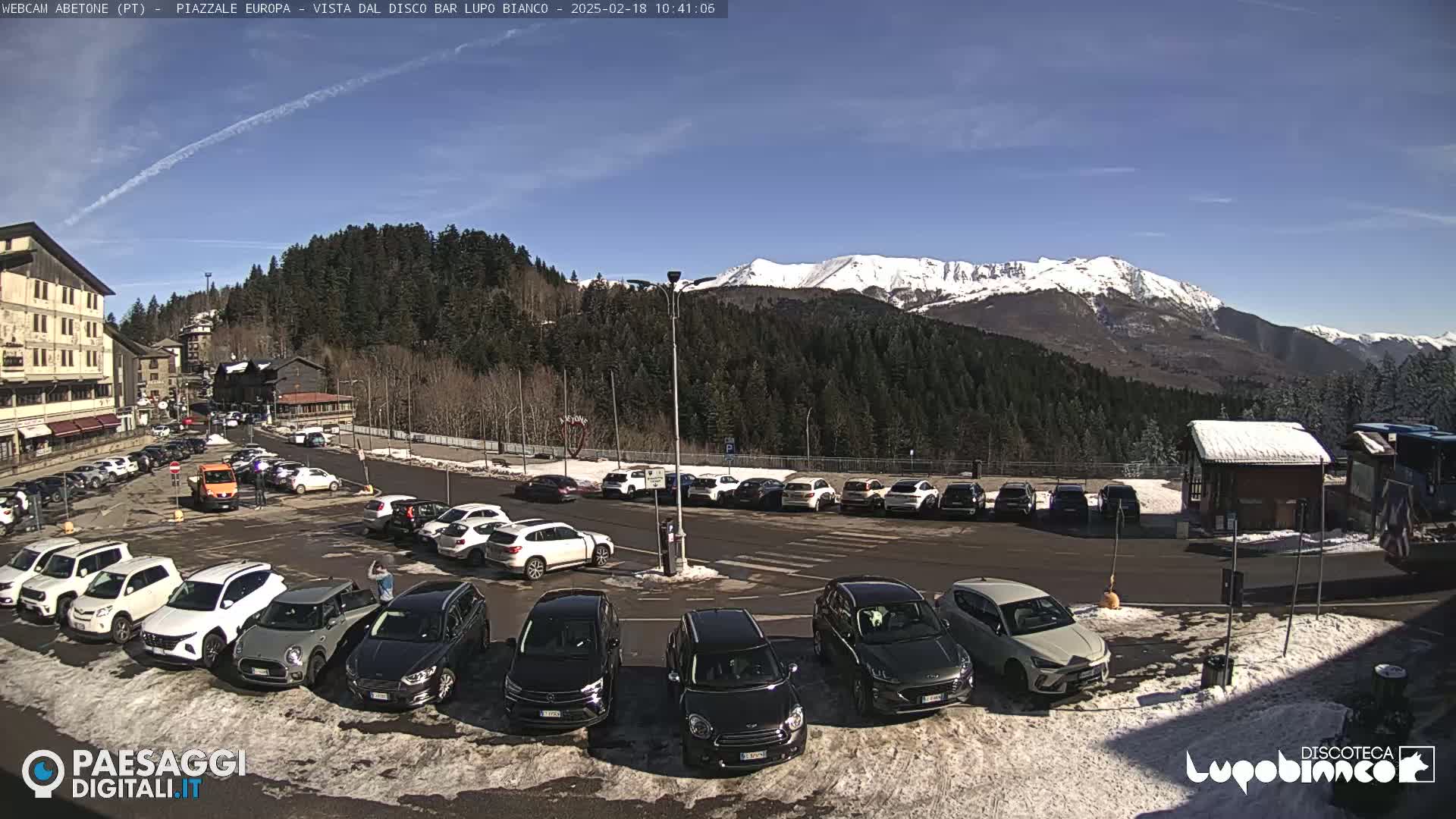 A sunny day reveals a parking lot filled with cars in front of a mountain range capped with snow.