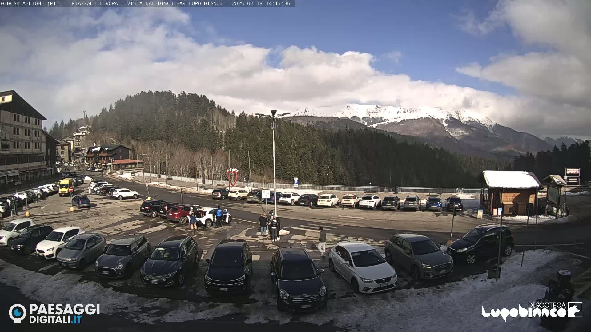 A partly sunny day in a mountain town shows a parking lot full of cars, a few people walking around, and snow-capped mountains in the background.