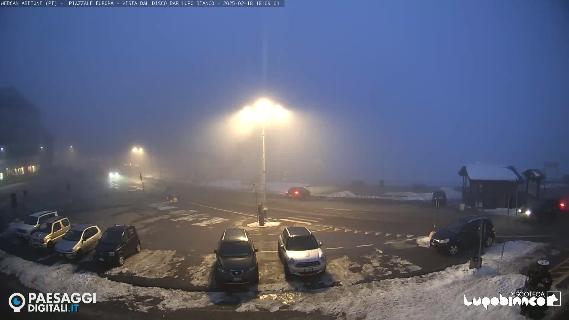 A foggy parking lot at night, with several cars parked and a few driving by, some snow patches on the ground, and streetlights illuminating the scene.