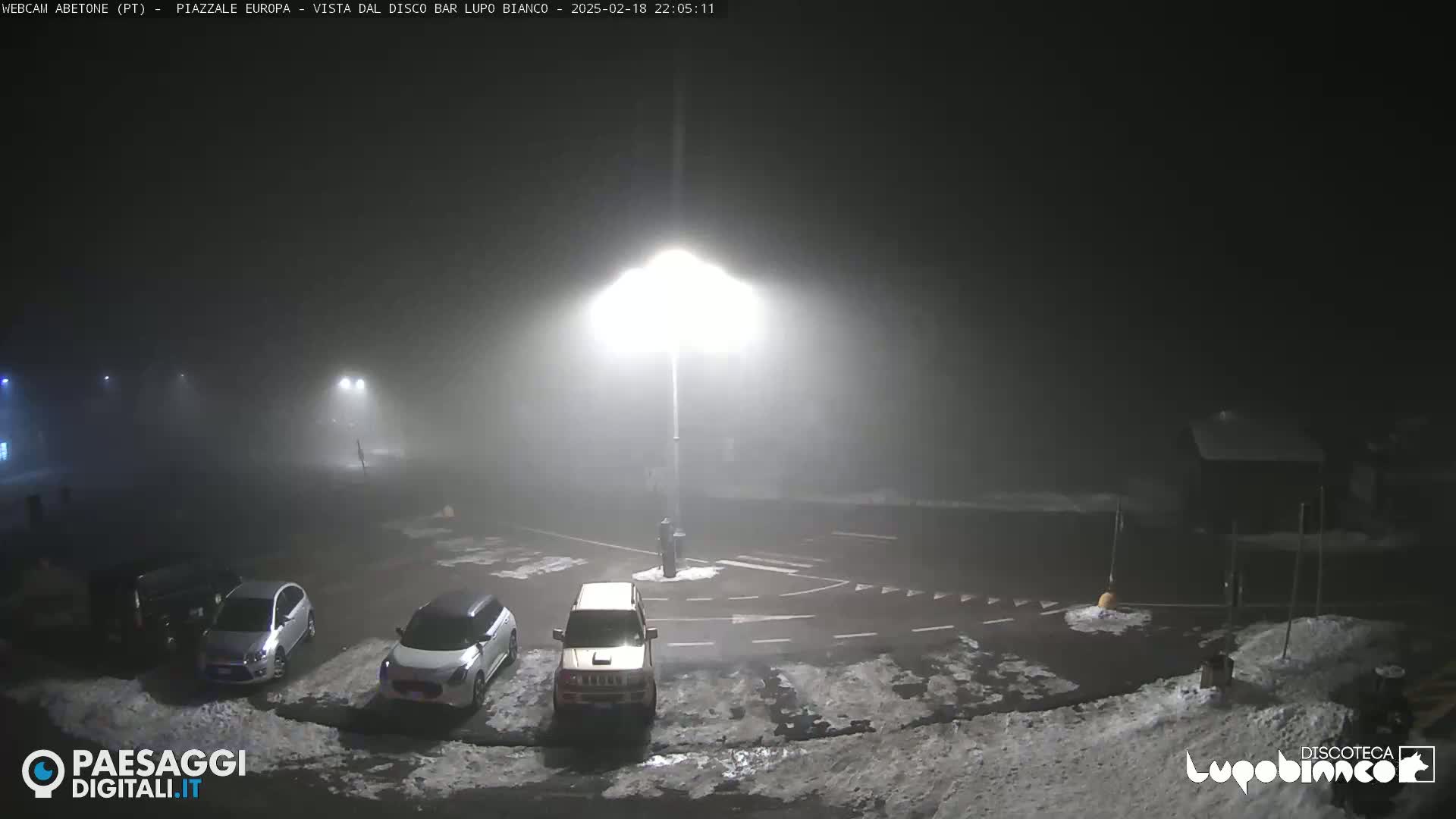 A foggy night scene shows a parking lot with several cars parked, illuminated by bright streetlights, with patches of snow on the ground.