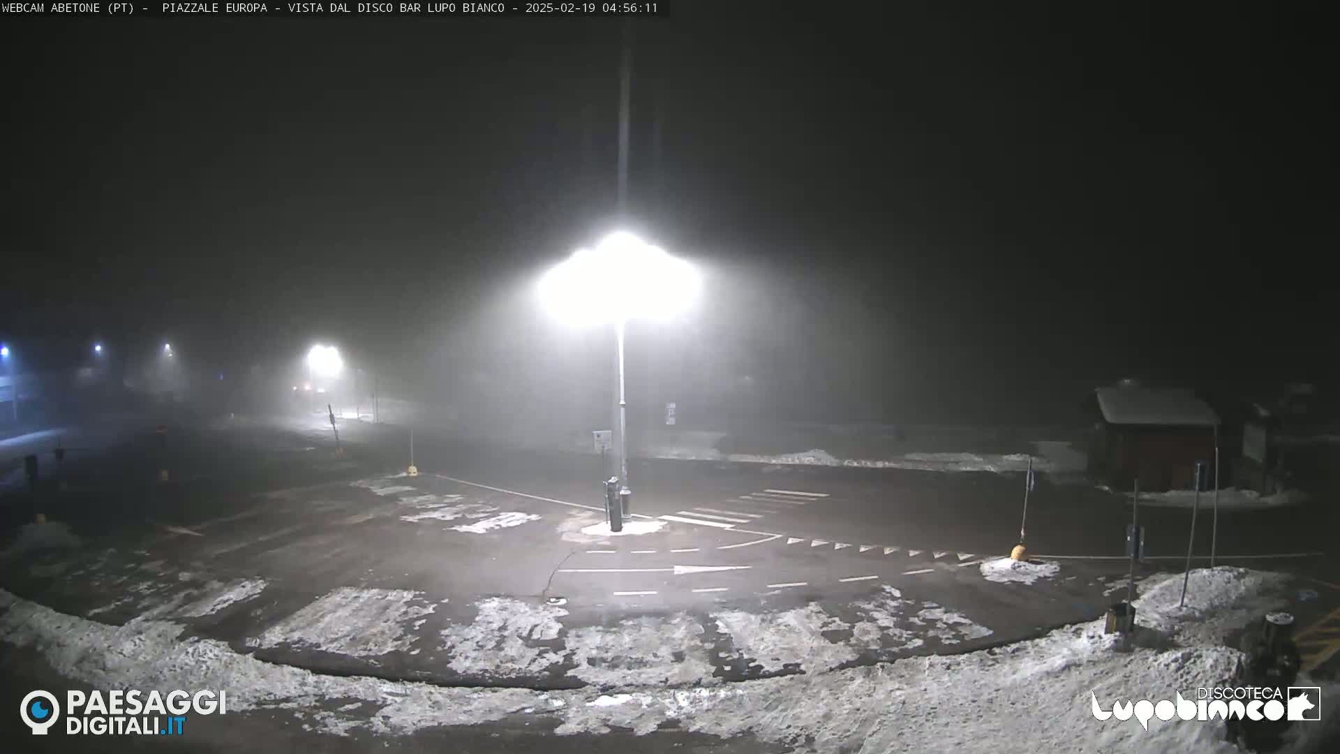 A nighttime, foggy parking lot with snow on the ground and streetlights illuminating the scene.