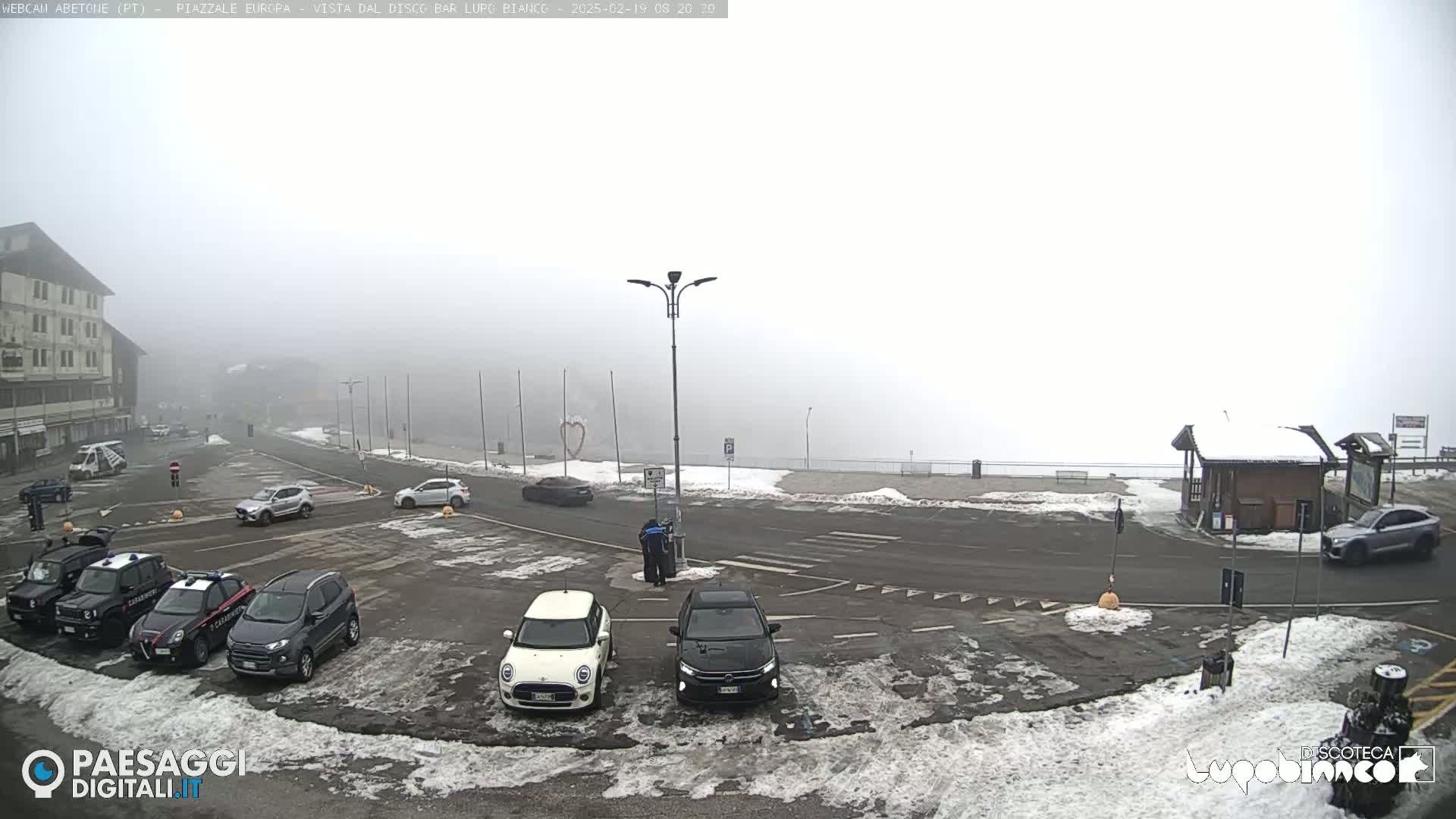 A foggy parking lot with several cars and a few buildings in the background, some patches of snow are on the ground.