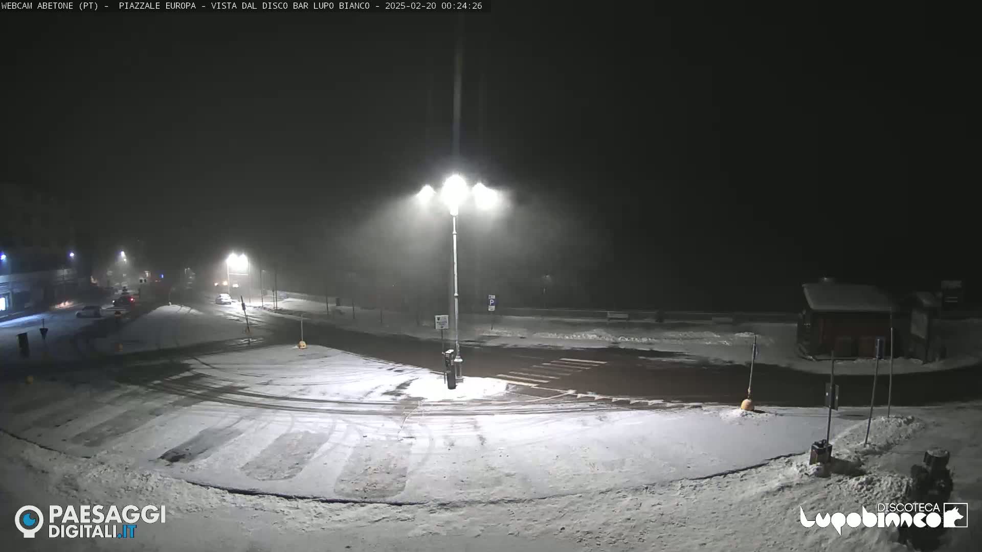 A snow-covered roundabout at night, illuminated by streetlights, with a few cars visible in the distance.