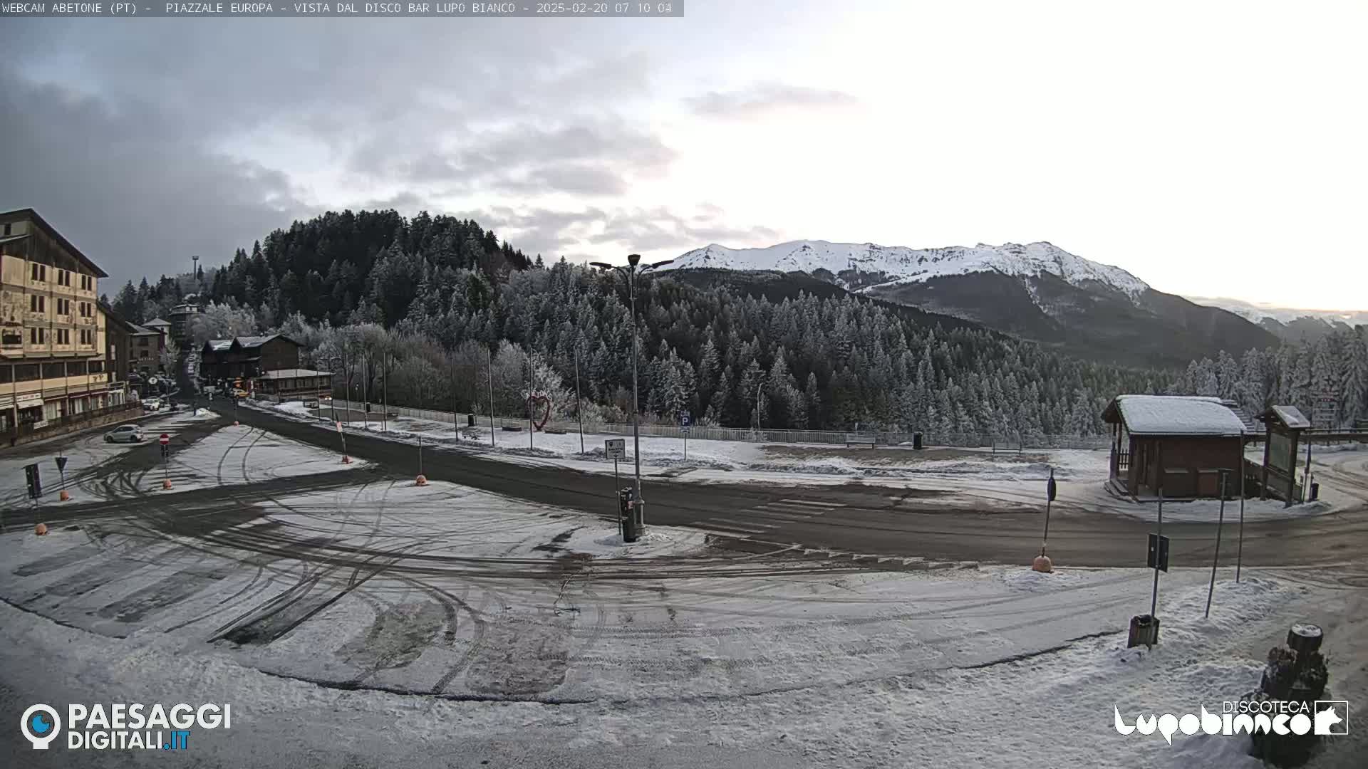 A snow-covered parking lot and roadway are situated in front of a building and a snow-dusted forest, with snow-capped mountains in the background under a cloudy sky.