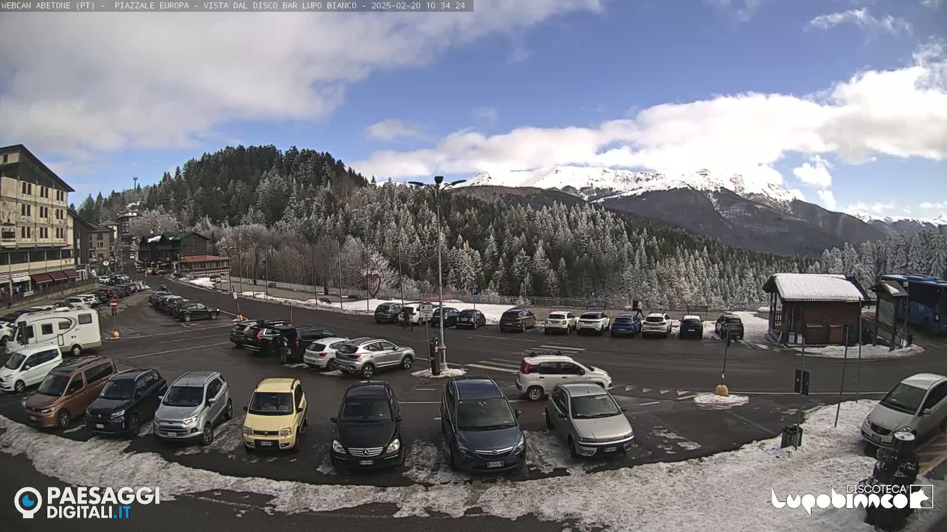 A snow-dusted parking lot filled with cars is situated in front of a snow-capped mountain range under a partly cloudy sky.