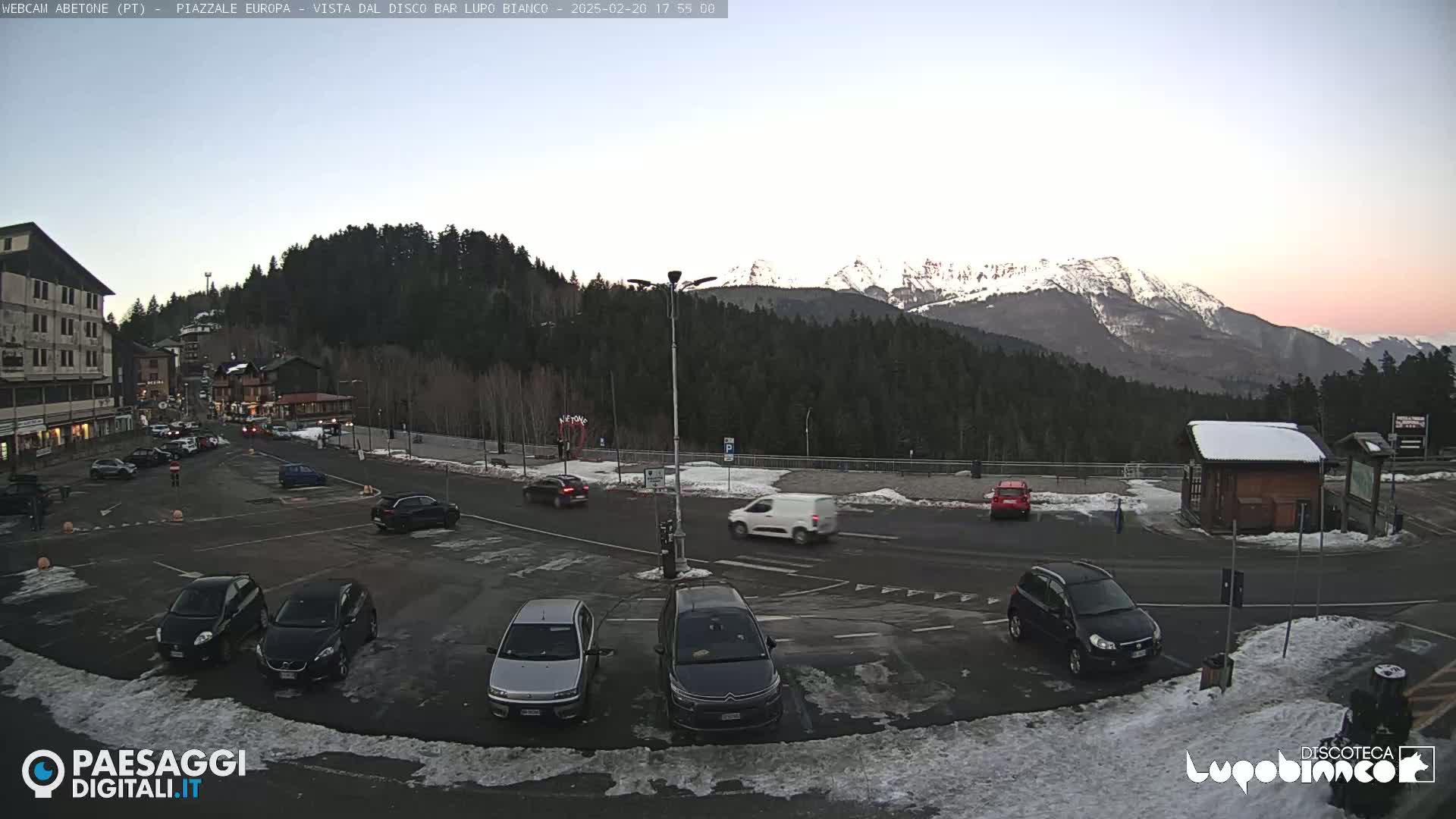 A parking lot with several cars is situated in front of buildings and a snowy mountain range under a twilight sky.