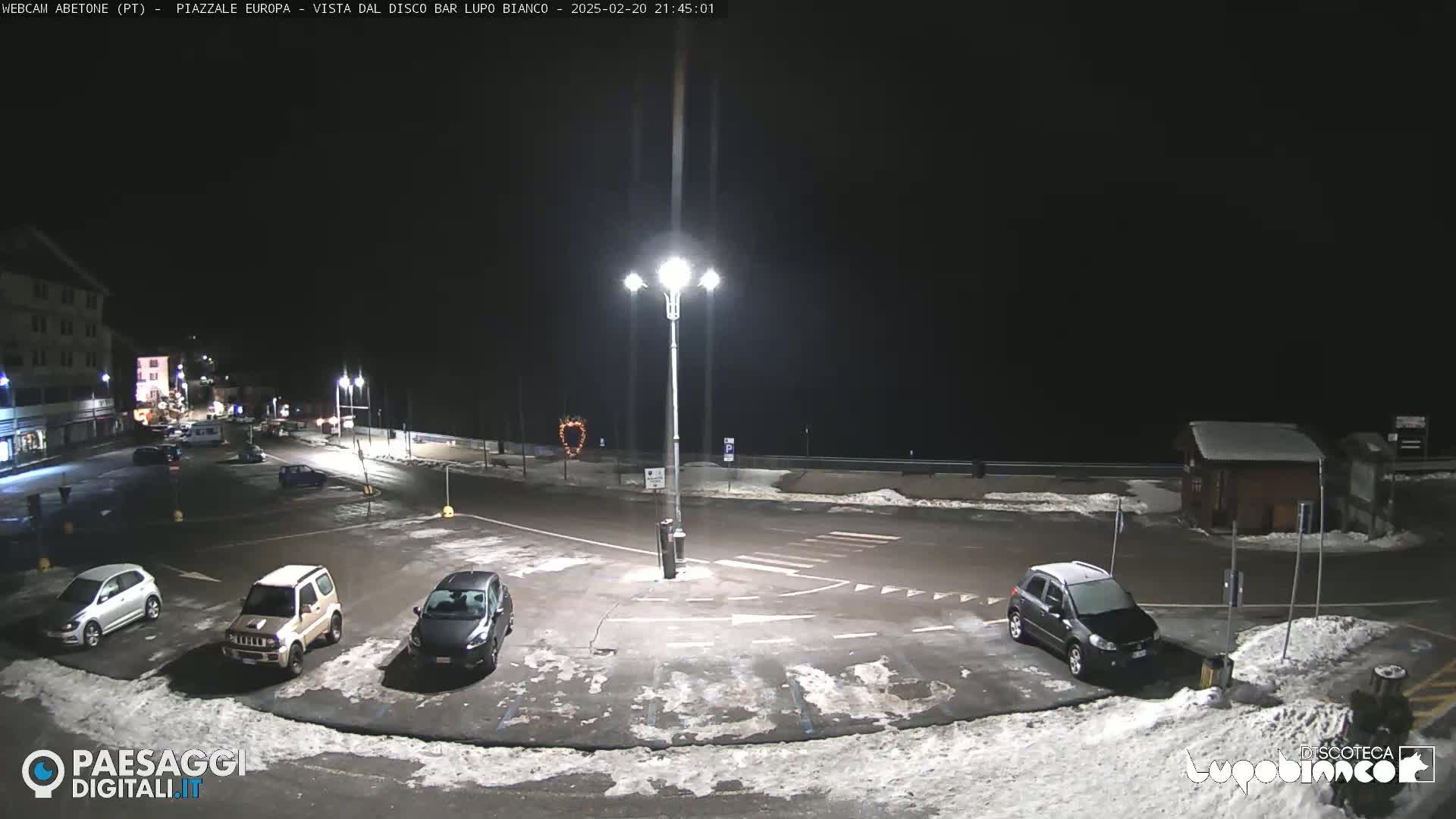 A nighttime view of a mostly empty parking lot with several cars, some snow on the ground, and a tall light pole in the center, under a dark, clear sky.