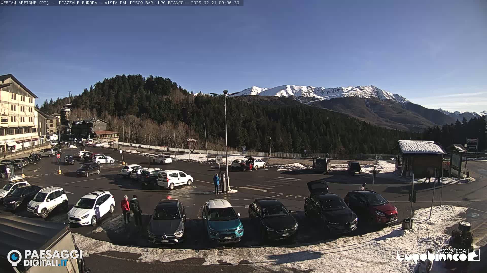 A sunny day in a mountain town parking lot with snow on the ground and snow-capped mountains in the background.