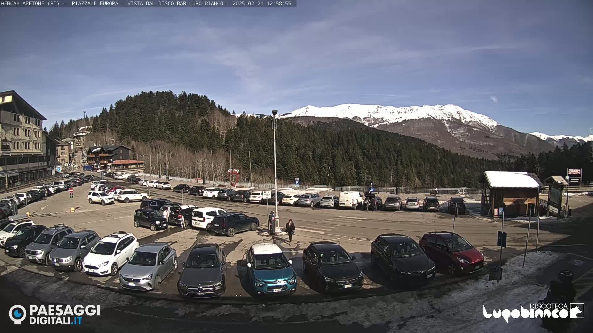 A sunny day in a mountain parking lot filled with cars, with snow-capped mountains in the background.