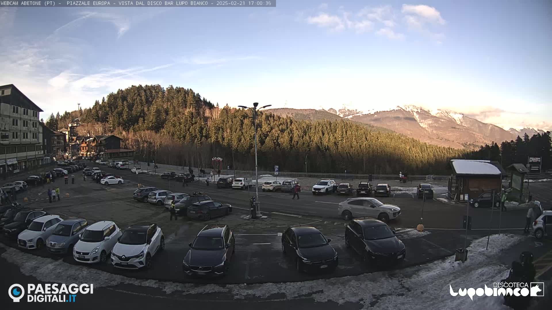 A mostly sunny day shows a large parking lot full of cars in a mountainous area with snow-capped peaks in the background.
