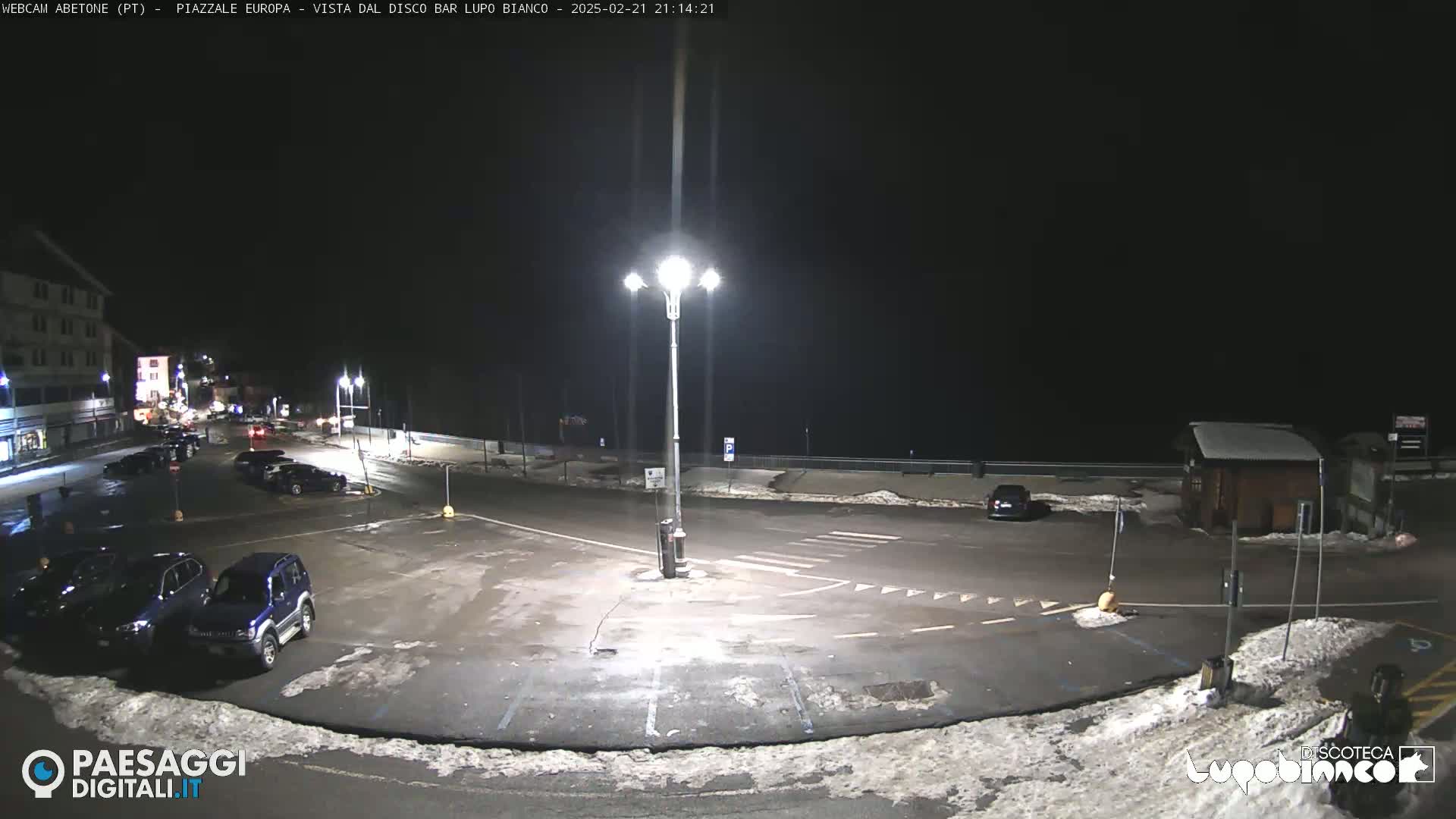 A nighttime view of a mostly empty parking lot with a few cars, illuminated by a tall streetlight, and some snow on the ground.