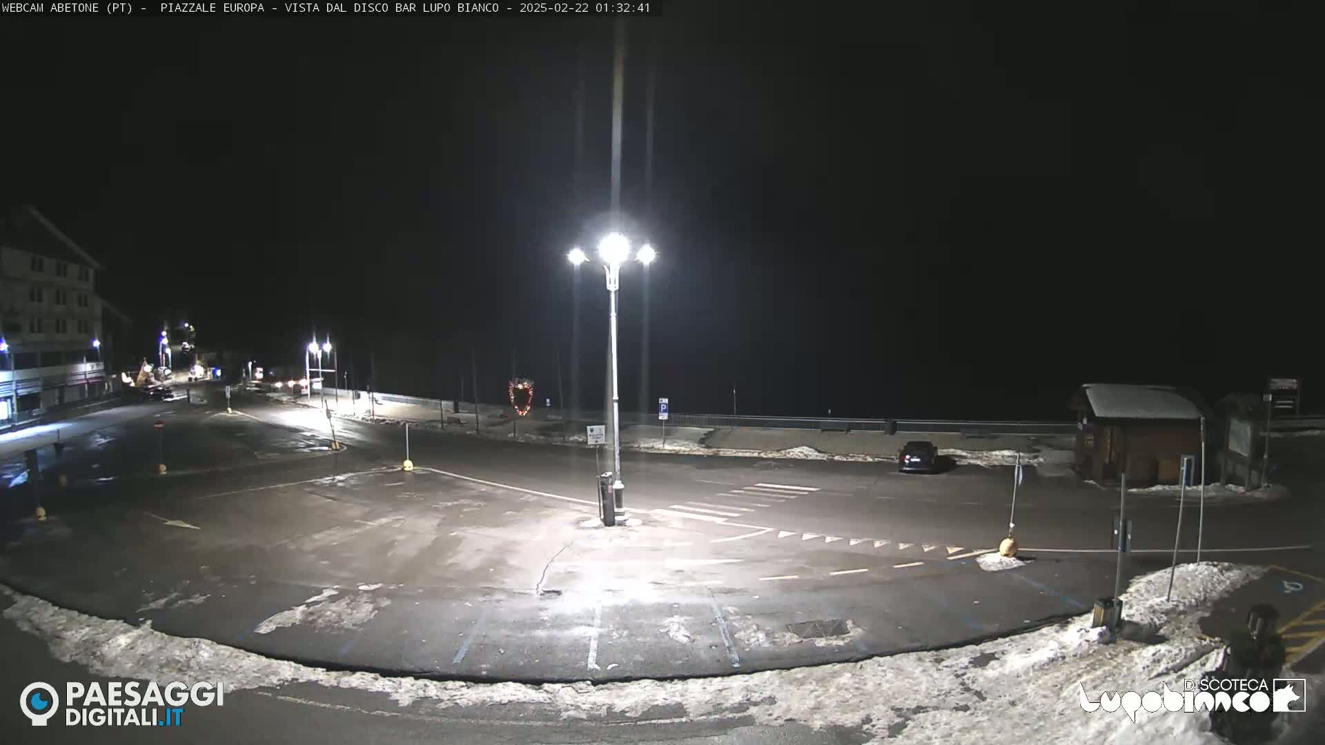 An empty, snow-dusted parking lot is illuminated by bright streetlights at night.