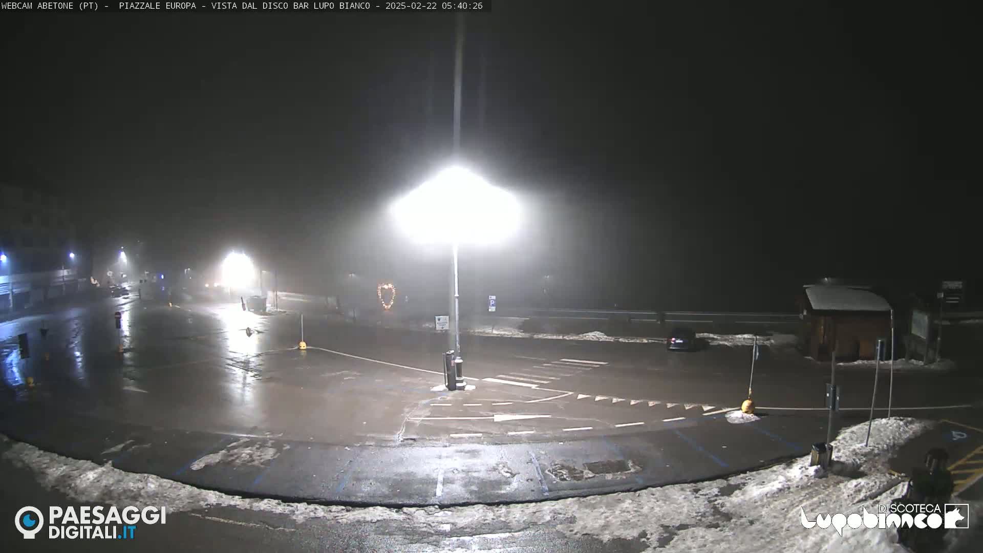 A foggy, nighttime scene shows a mostly empty parking lot with a few cars and some snow on the ground, illuminated by bright streetlights.