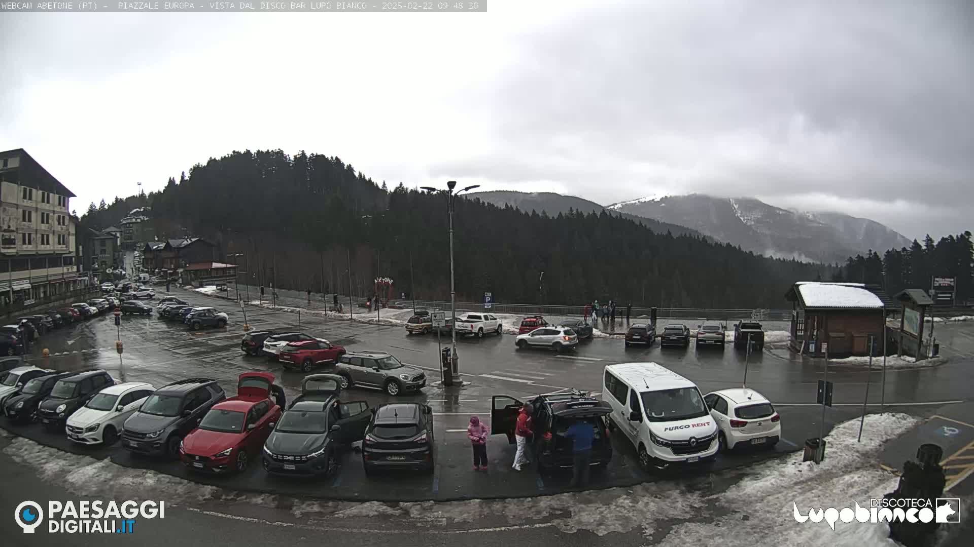 A wet parking lot filled with cars is situated in front of a building, with a mountainous backdrop partially obscured by clouds.