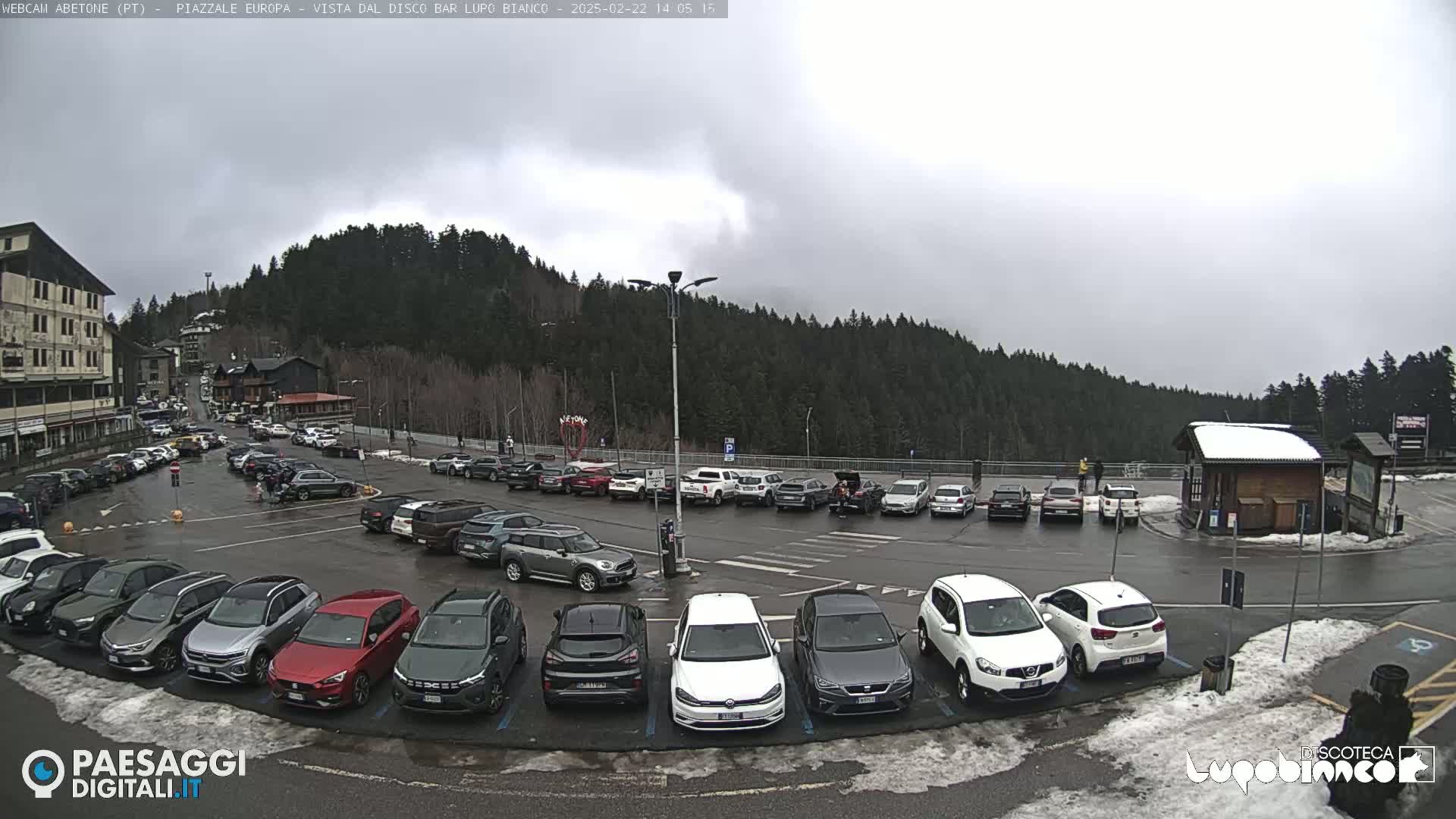 A parking lot filled with cars is situated in front of a forested mountain under an overcast sky with some snow on the ground.