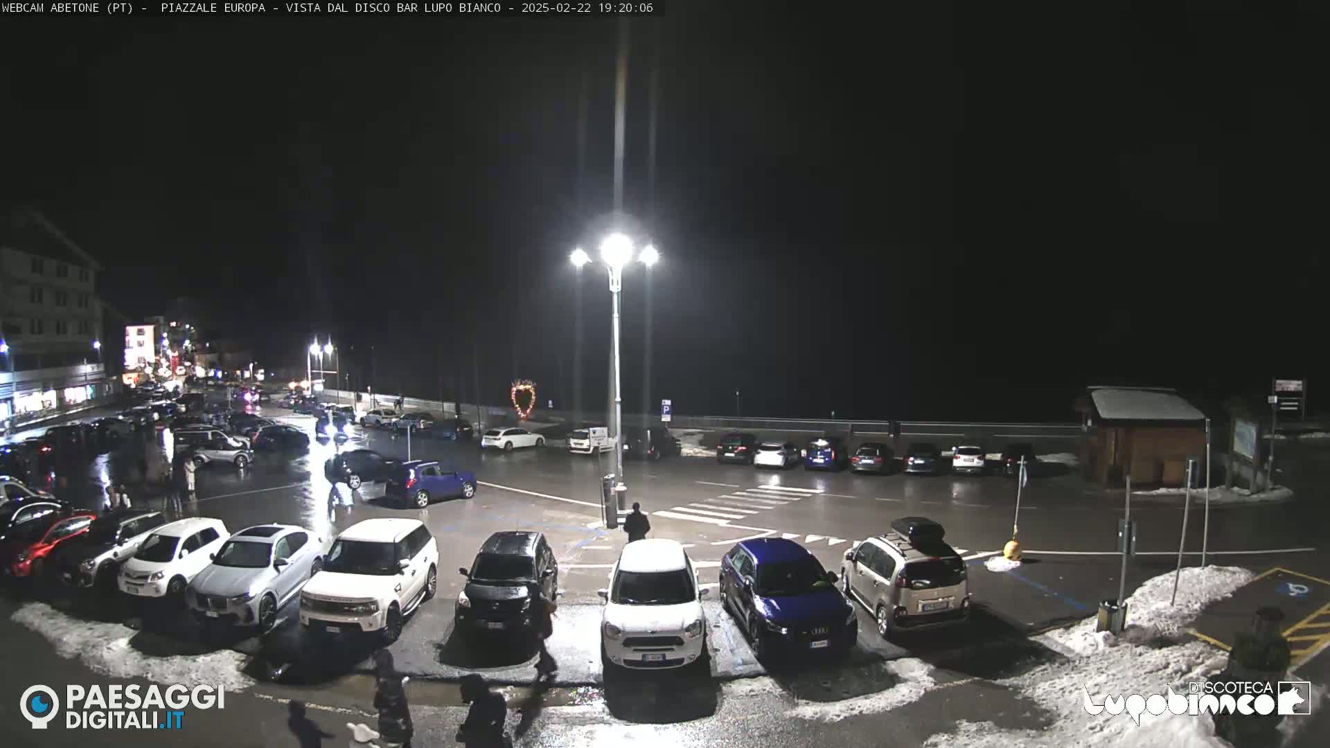 A nighttime, wet parking lot with numerous cars is illuminated by streetlights, with some snow visible on the ground.