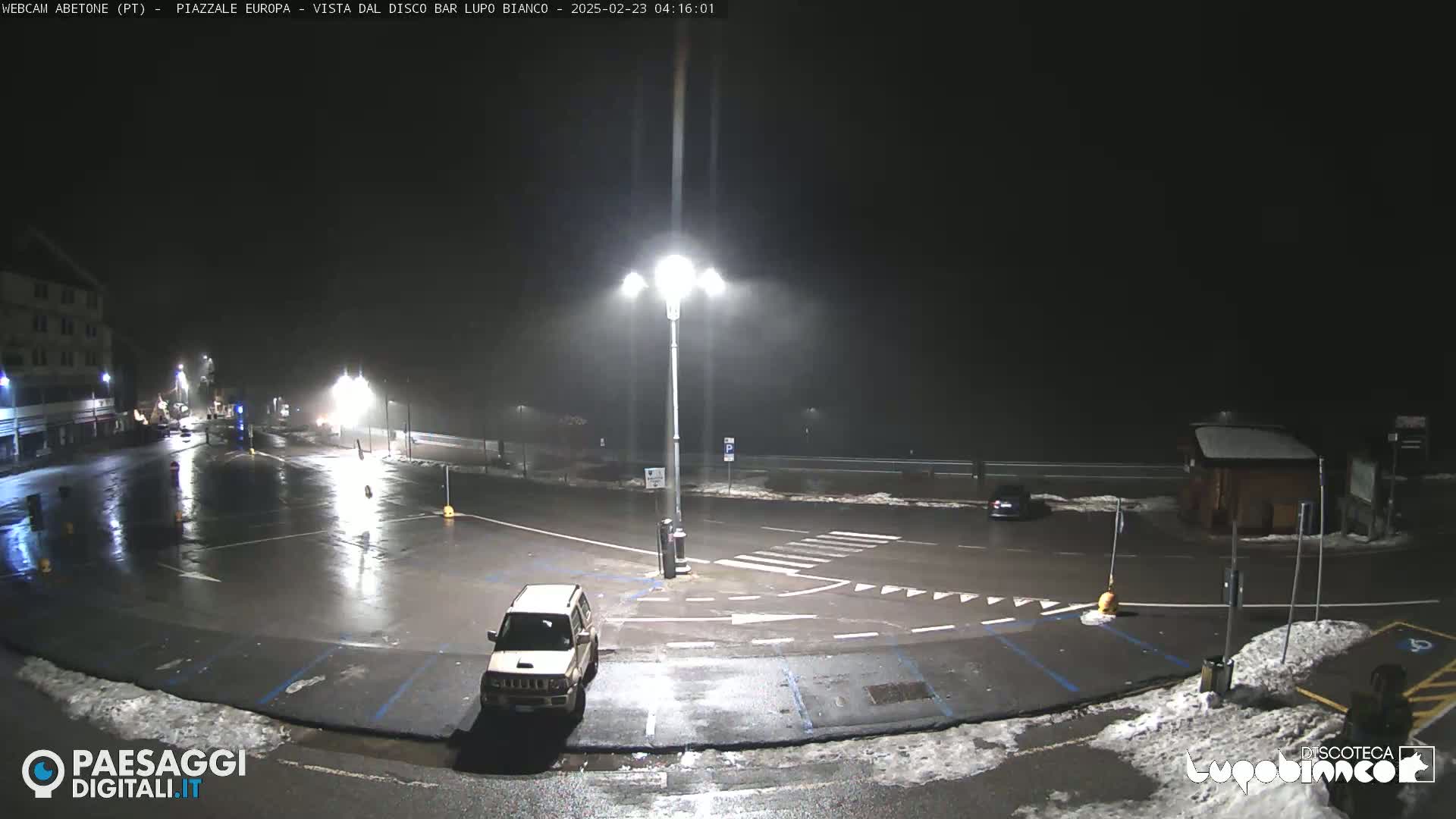 A rain-slicked, empty parking lot at night, illuminated by streetlights, with a small SUV parked near a crosswalk, and a few buildings in the distance.