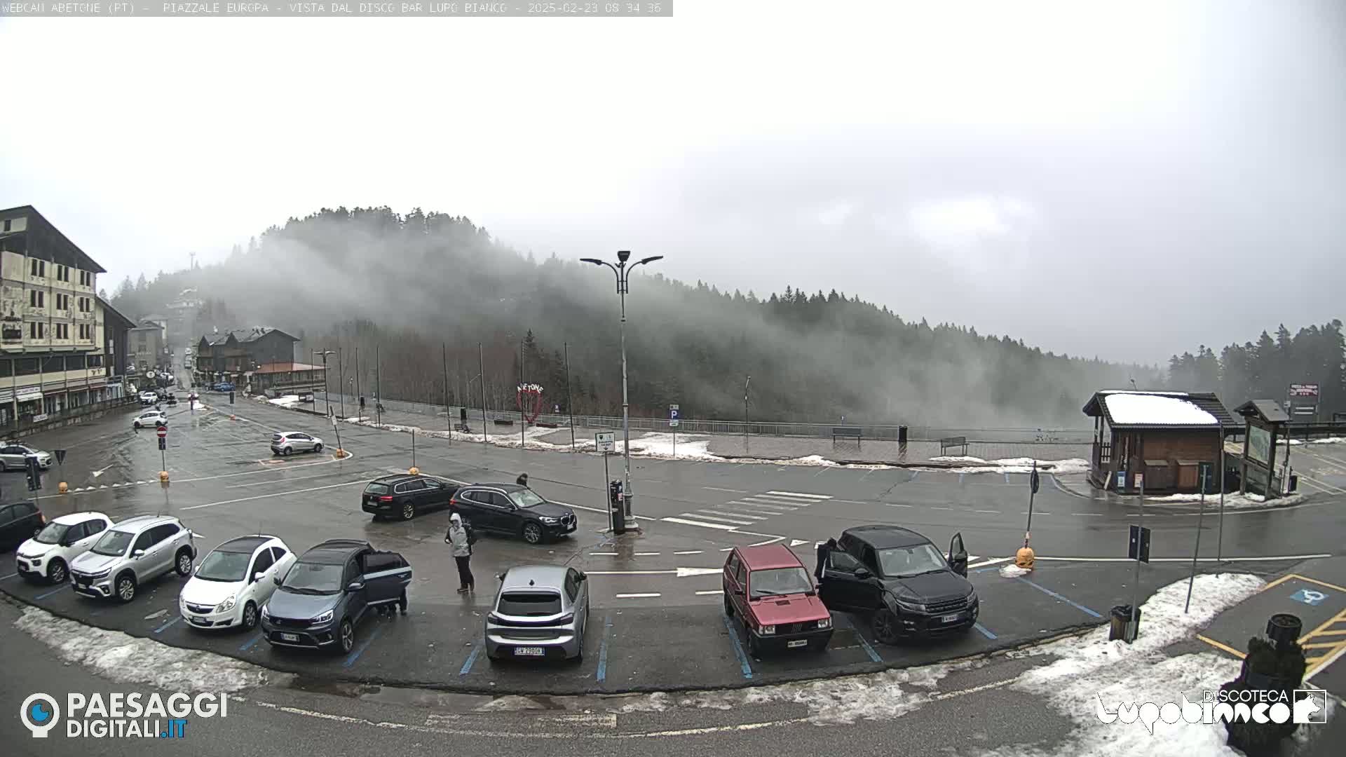 A foggy, wet parking lot with several cars and a few people, situated in front of a building and a hill covered in low clouds and trees.