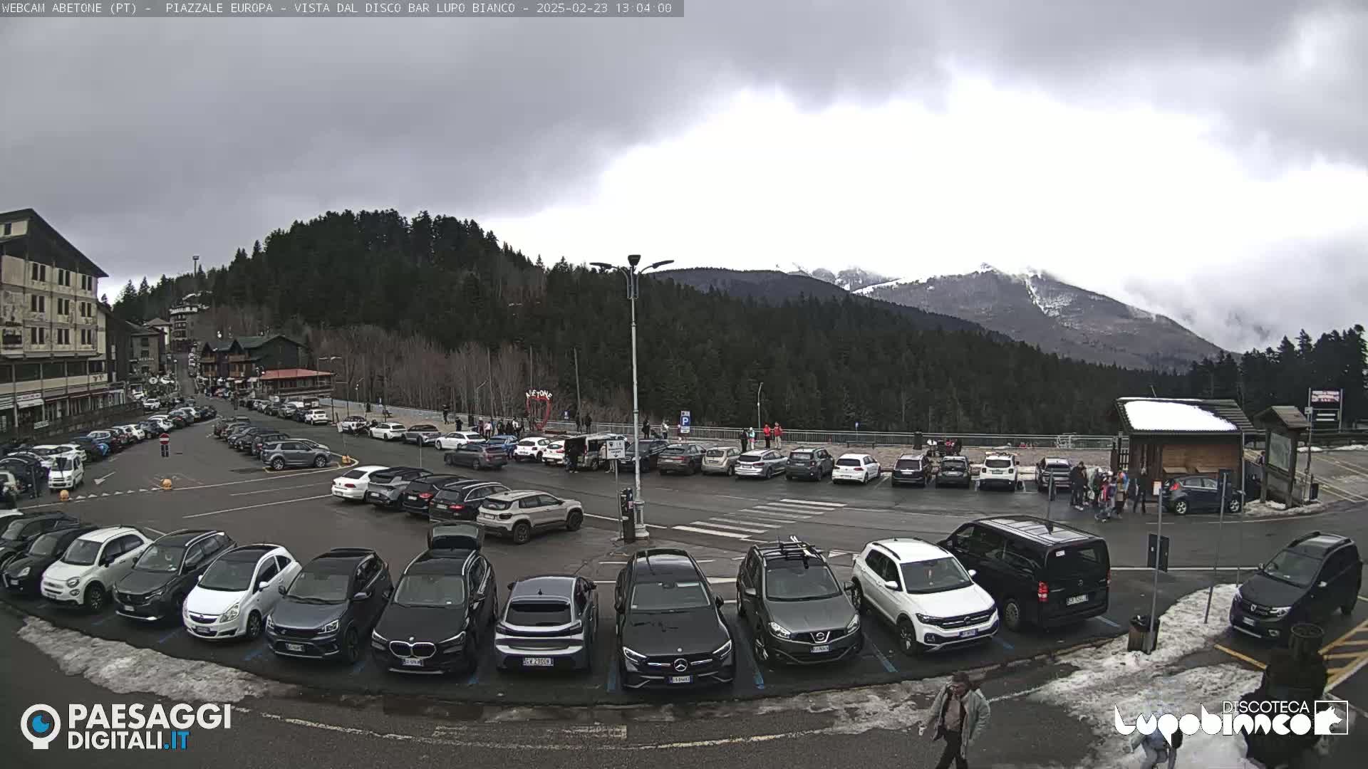 A mostly cloudy day reveals a full parking lot surrounded by buildings and evergreen trees, with snow-capped mountains in the distance.