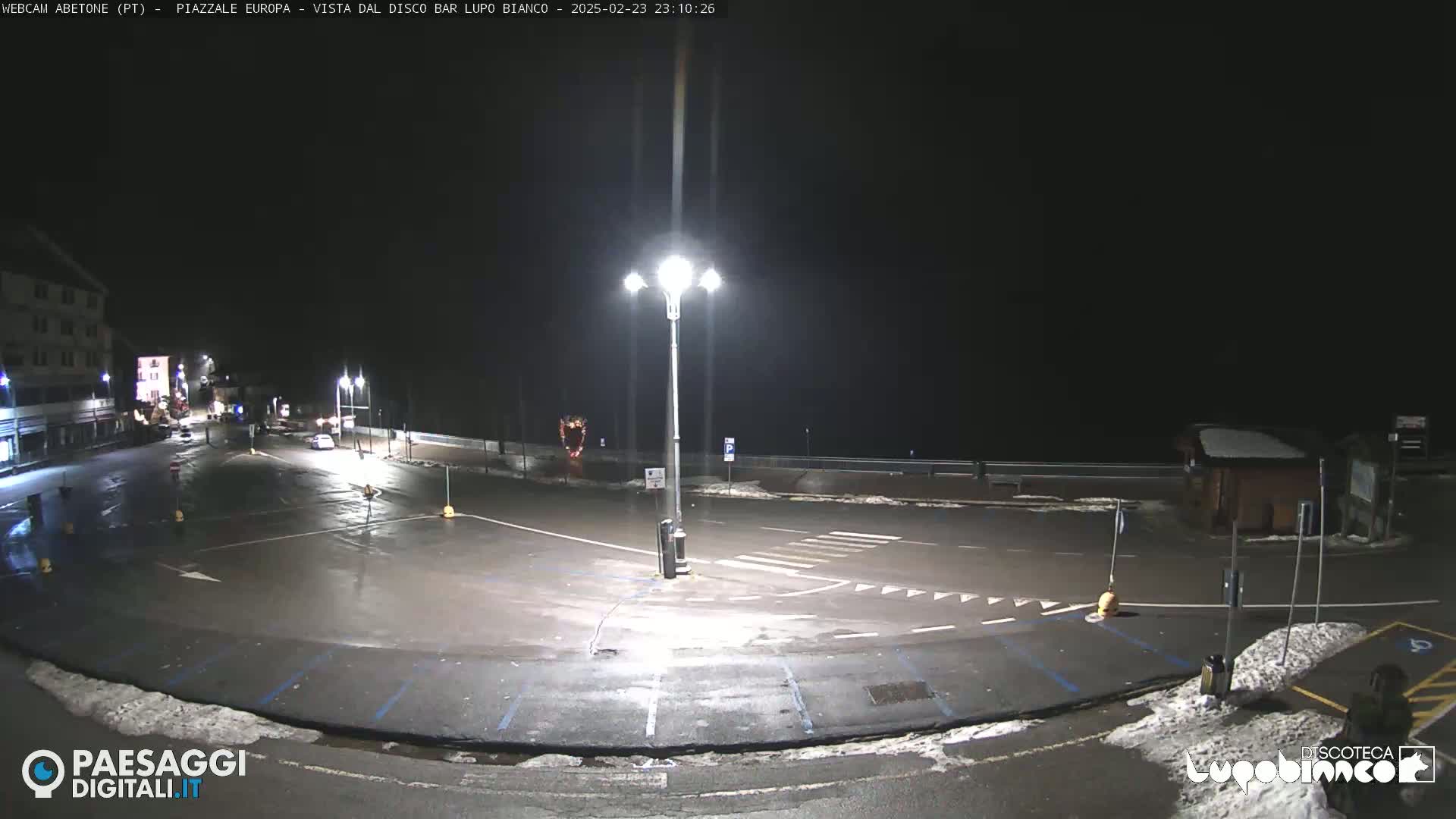 A mostly empty, wet parking lot is illuminated by bright streetlights at night, with some snow visible on the ground.
