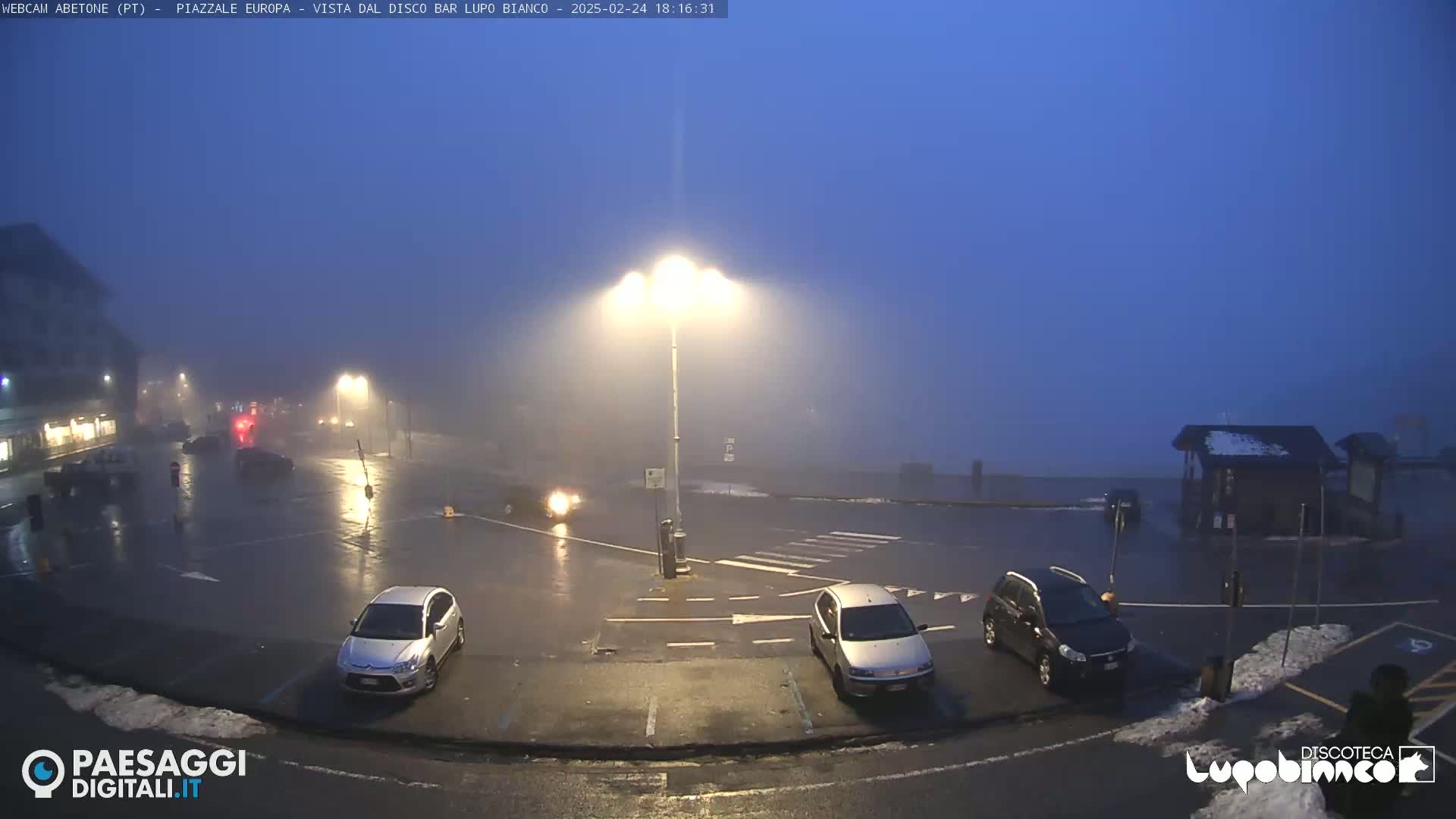 A foggy, wet parking lot at night is illuminated by streetlights, with several cars parked and a few driving through.