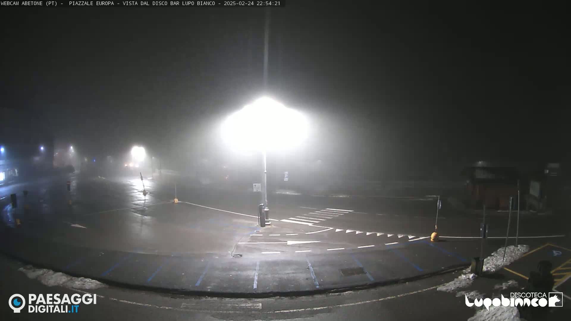 A foggy, nighttime view of an empty parking lot and roadway intersection, illuminated by bright streetlights.