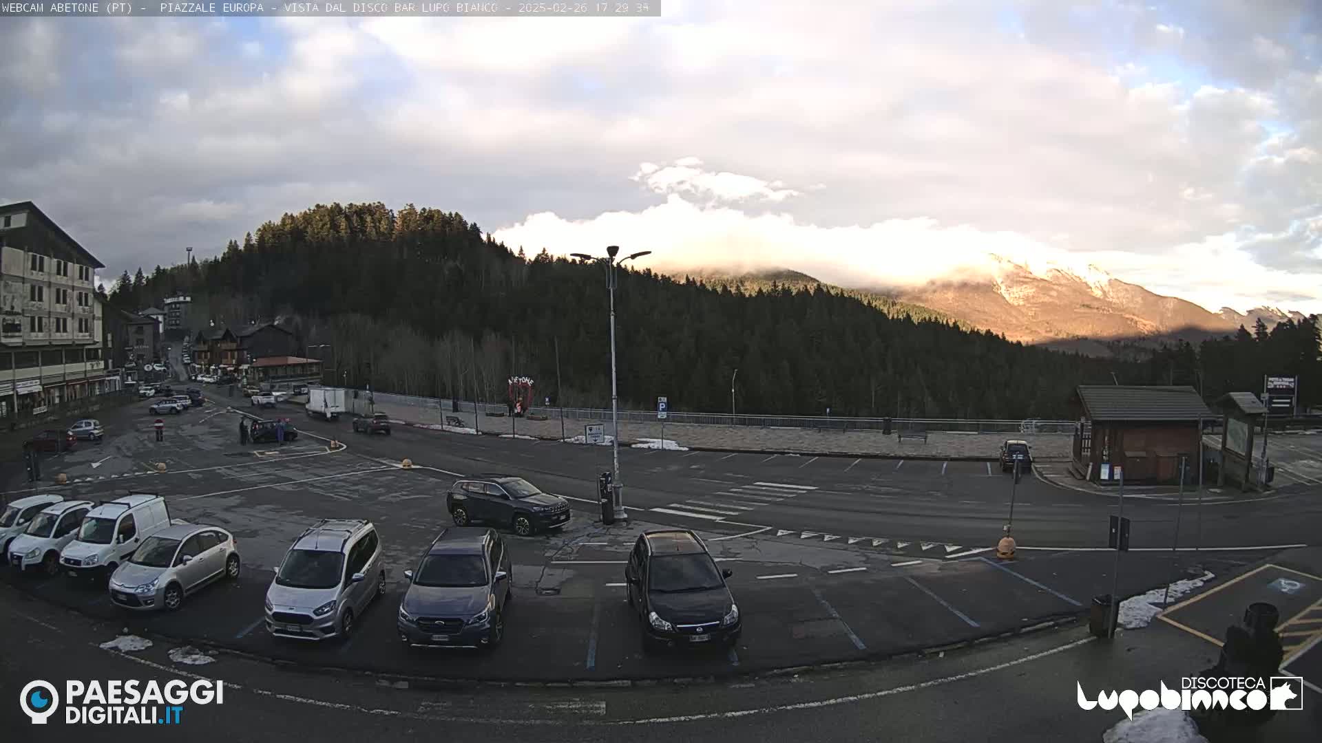 A mostly cloudy day reveals a mountain range partially shrouded in mist, overlooking a parking lot filled with cars near a building complex.