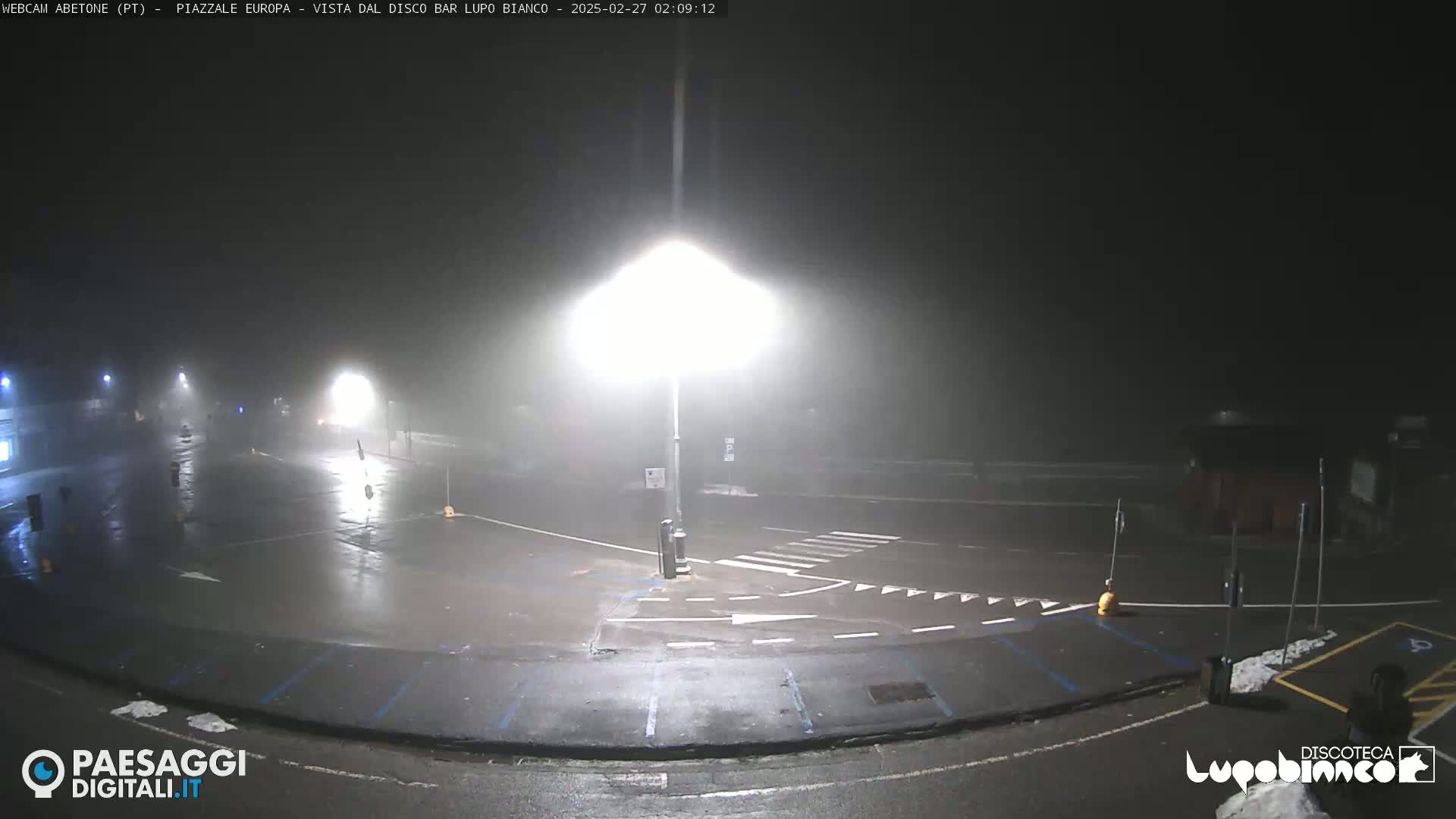 A foggy, wet nighttime scene shows an empty parking lot and road intersection illuminated by bright streetlights.