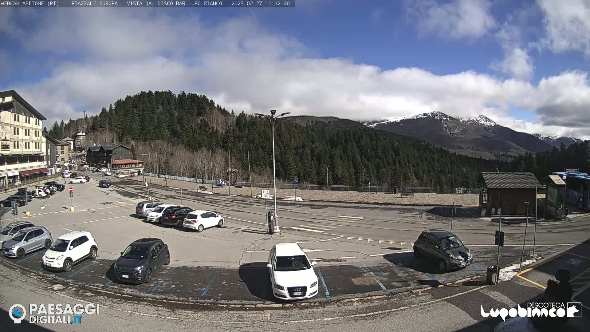 A partly cloudy sunny day reveals a parking lot full of cars in front of buildings and a mountain range with some snow-capped peaks in the background.