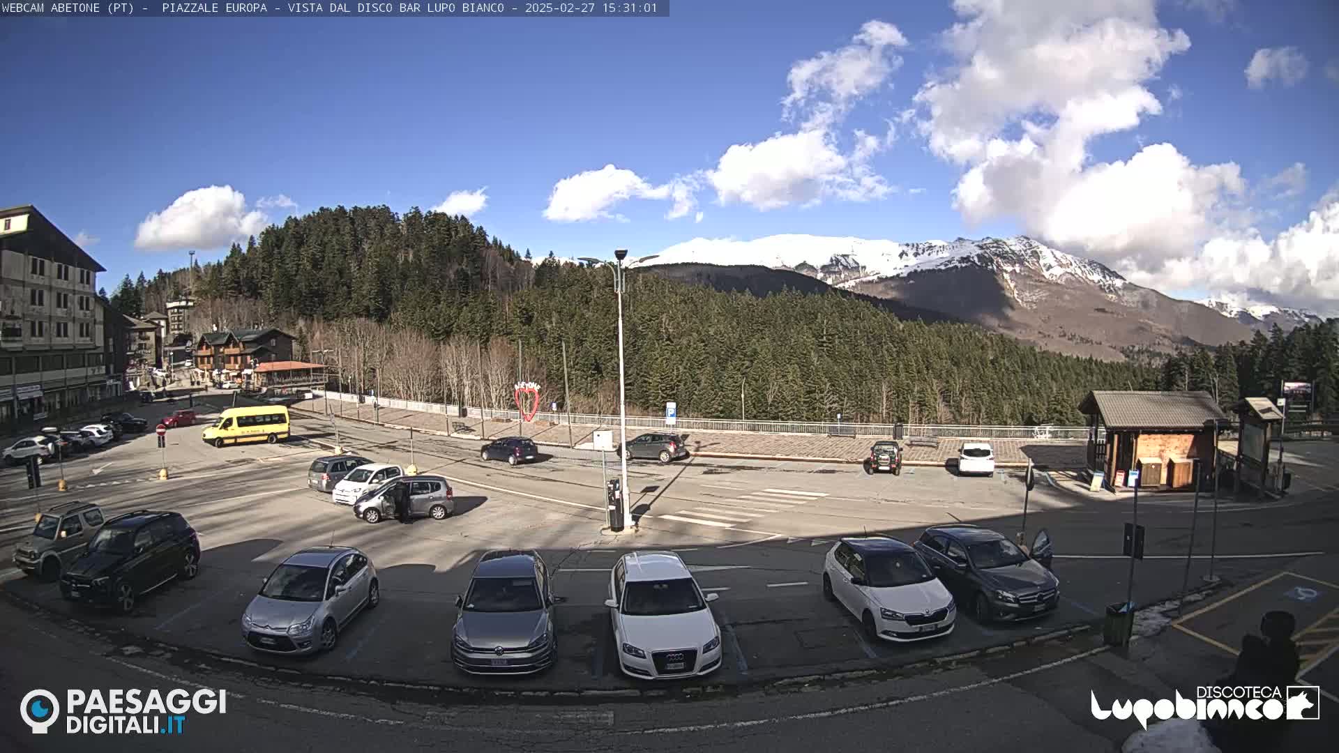 A mostly sunny parking lot with several cars and a bus, situated in front of a building and a forested mountain range with snow-capped peaks in the background.