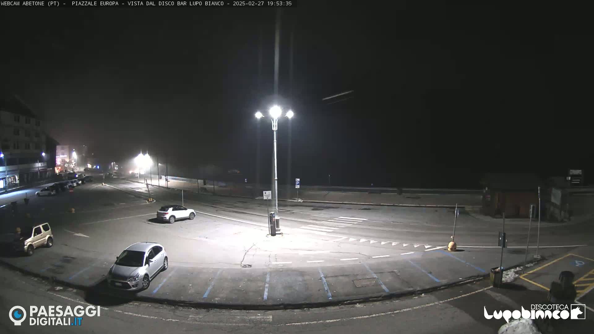 A nighttime, foggy view of a mostly empty parking lot with a few cars, illuminated by streetlights.