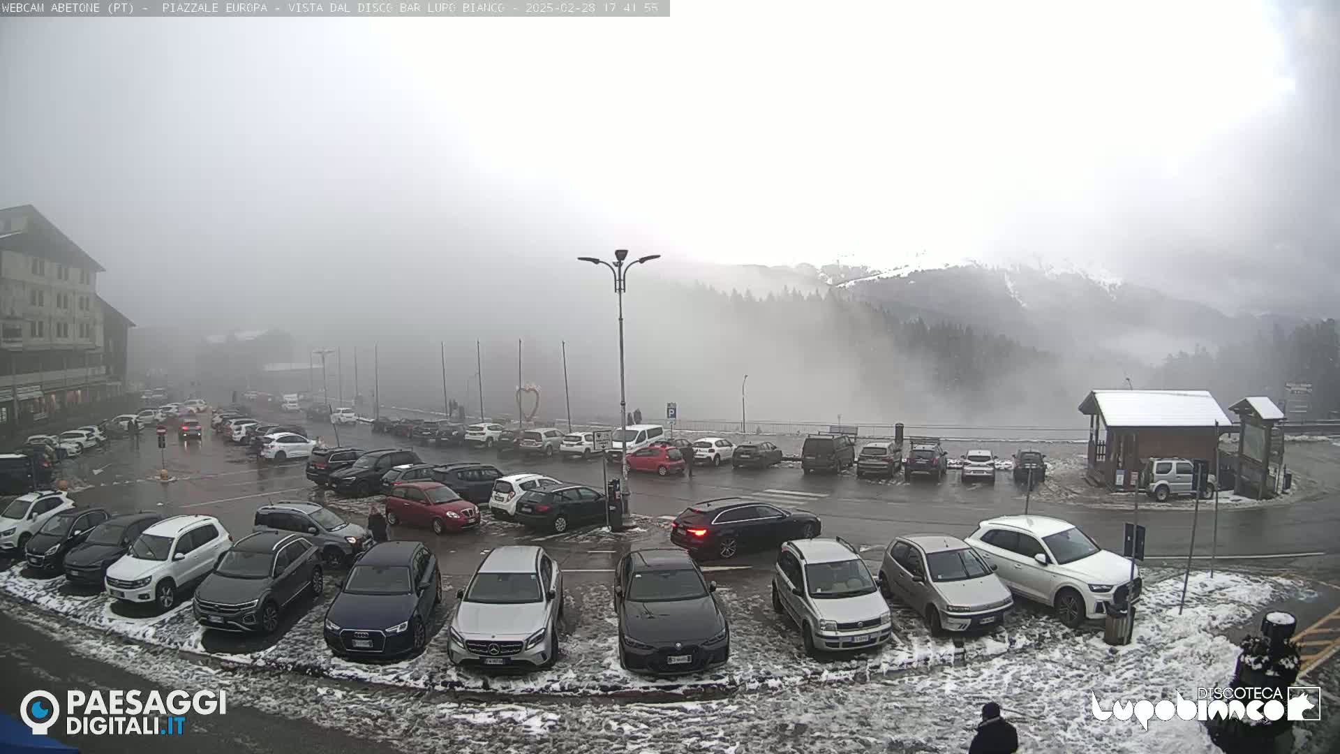 A parking lot filled with cars is shown, situated in front of a foggy mountain landscape with a light dusting of snow on the ground.